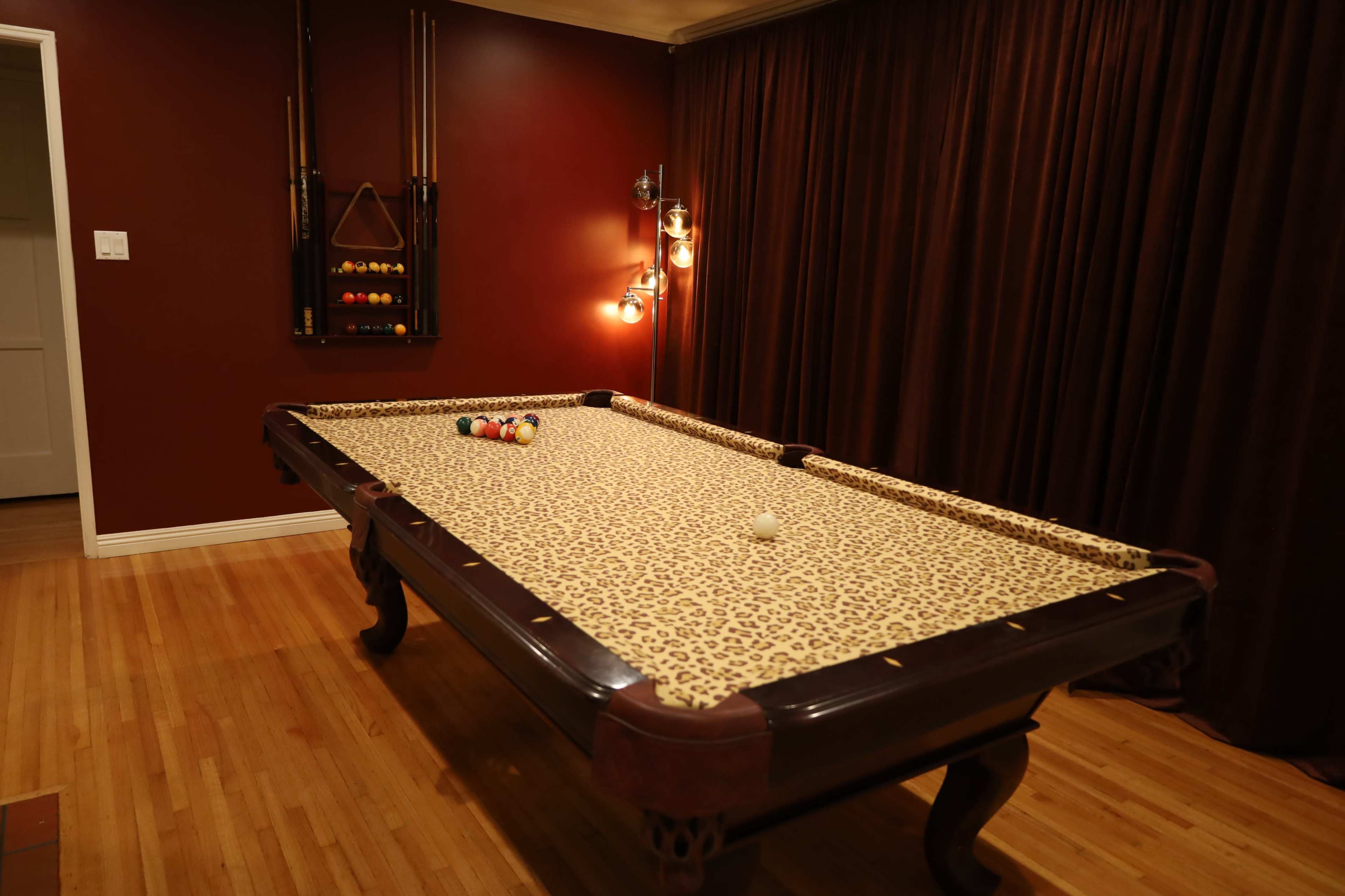 The image shows a billiard table with a leopard print felt located in a room with dark red walls and wooden flooring.