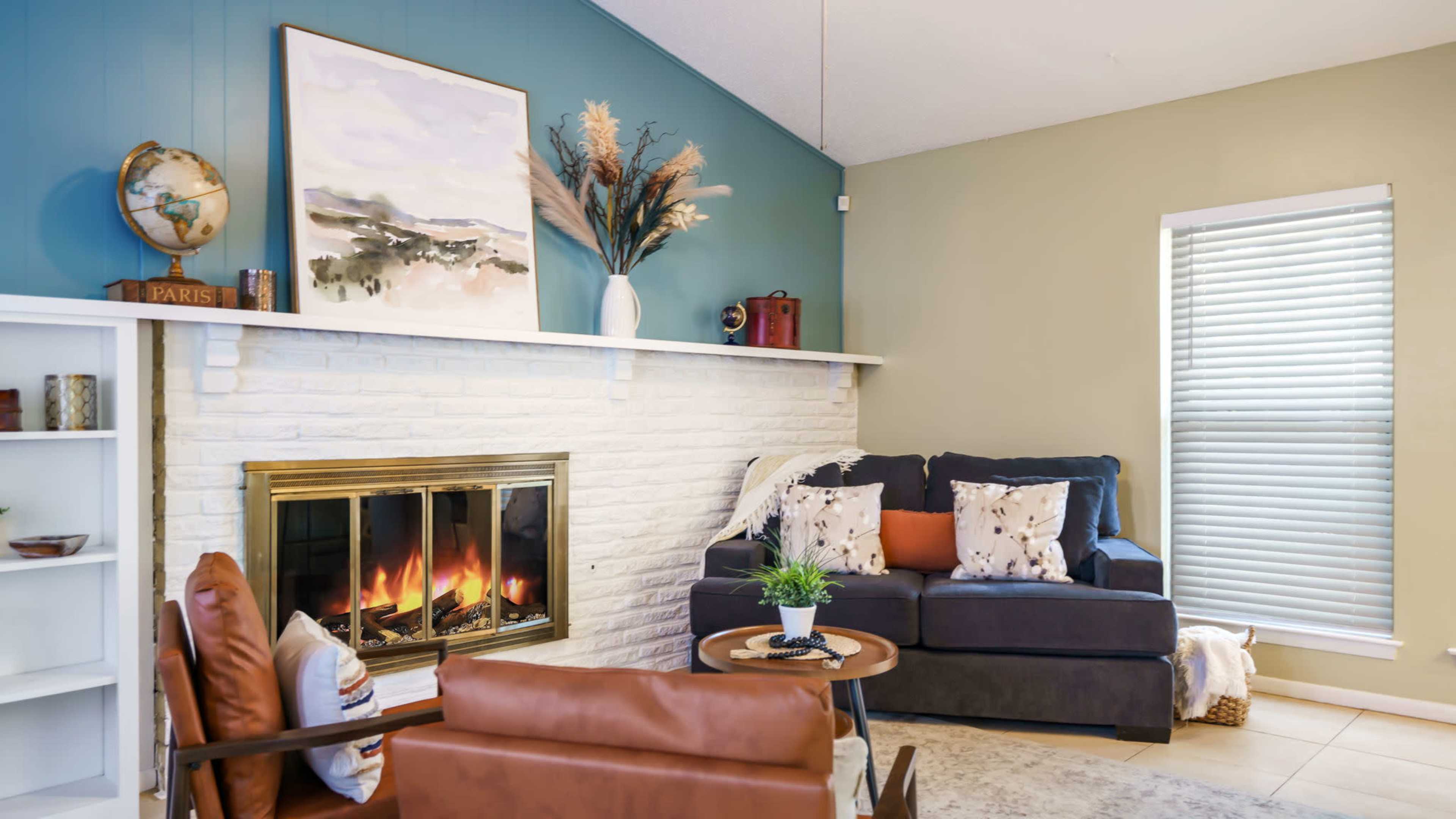 The living room features a brick fireplace, a blue accent wall, a dark sofa with patterned cushions, and a small coffee table beside brown leather chairs.
