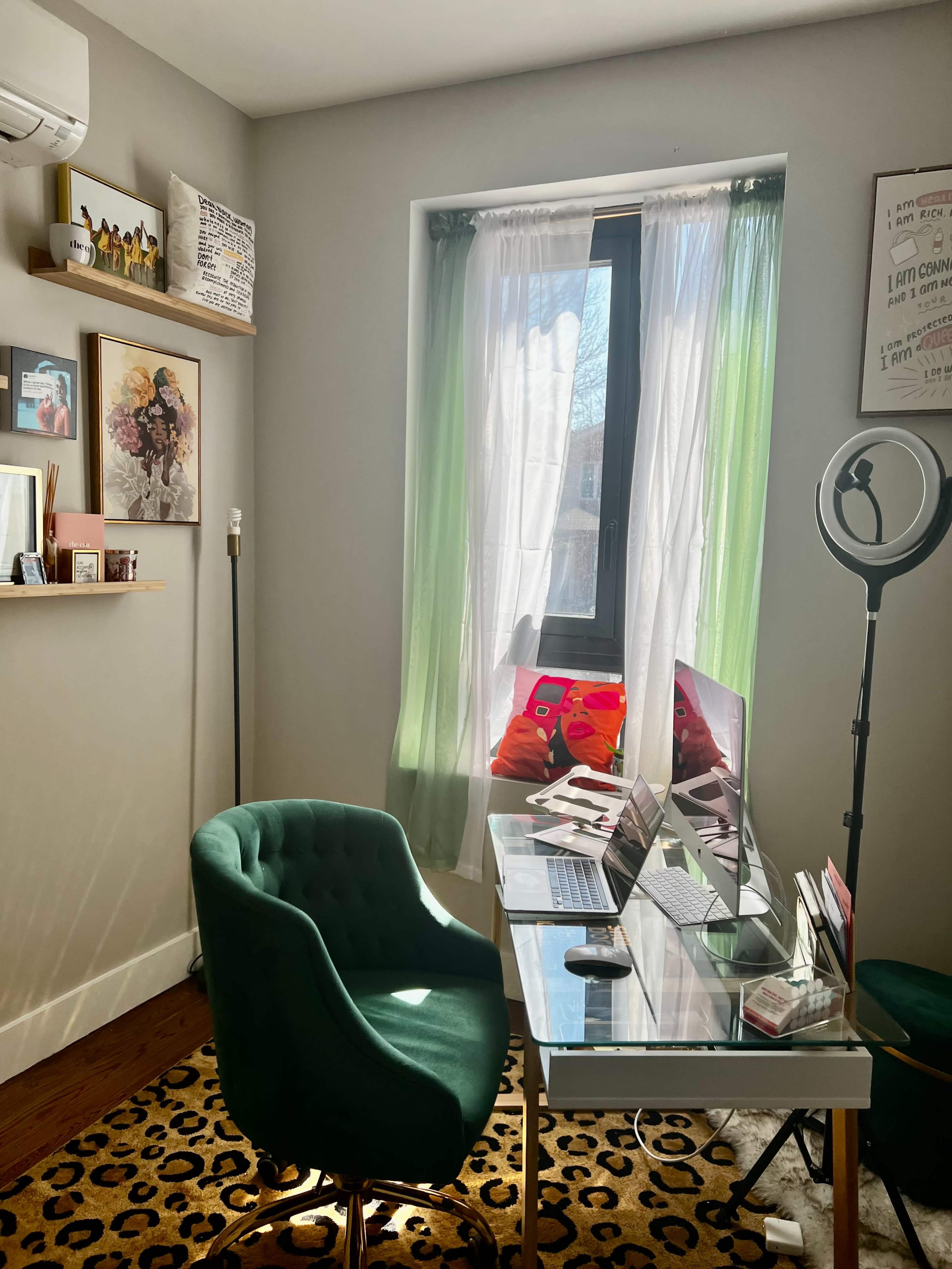 The image shows a neatly organized home office with a glass desk, a green chair, and natural light coming through a window with sheer curtains.