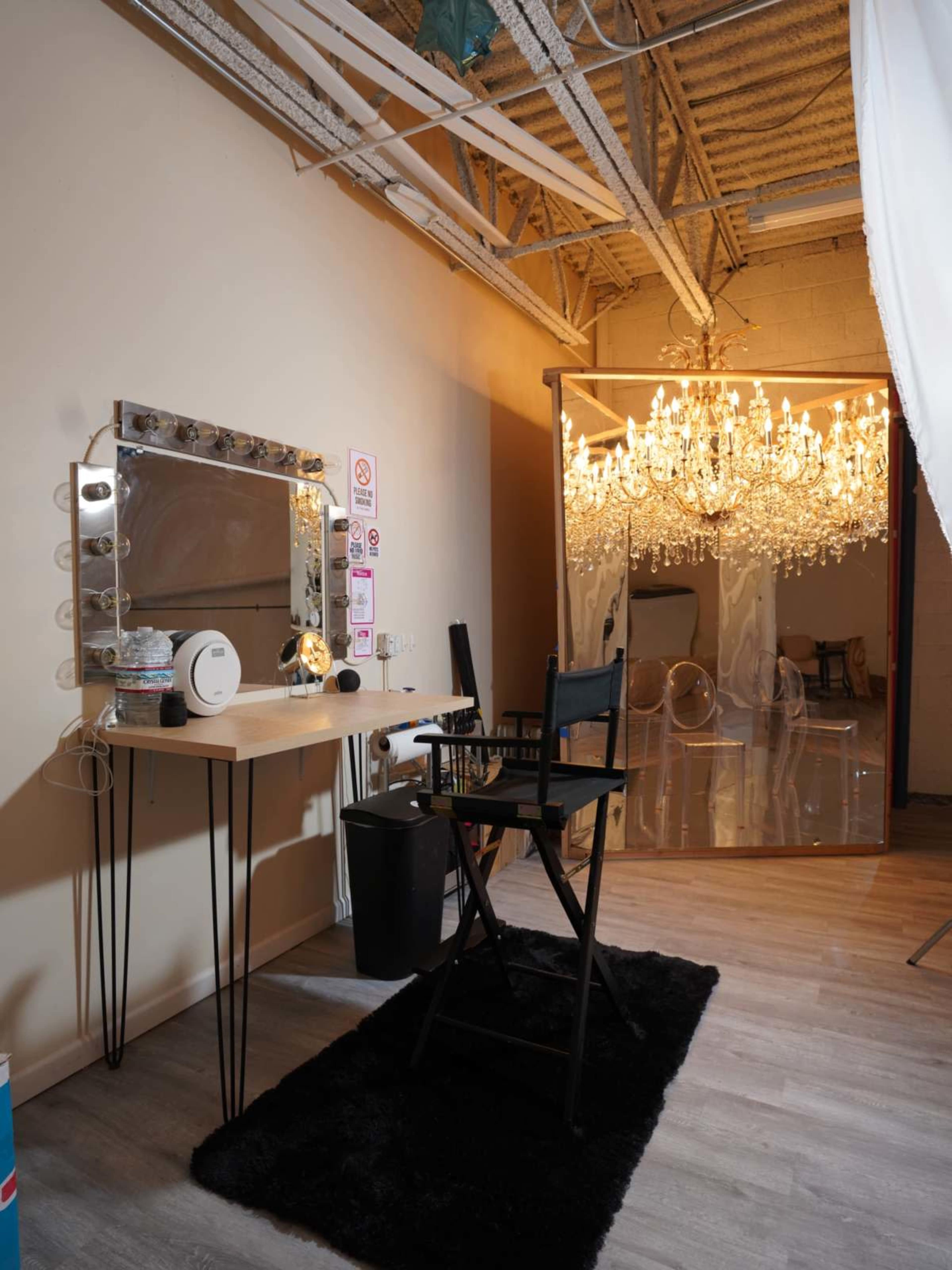 The image shows a well-lit makeup station with a large mirror and illuminated vanity lights, accompanied by a director's chair and a chandelier in the background.