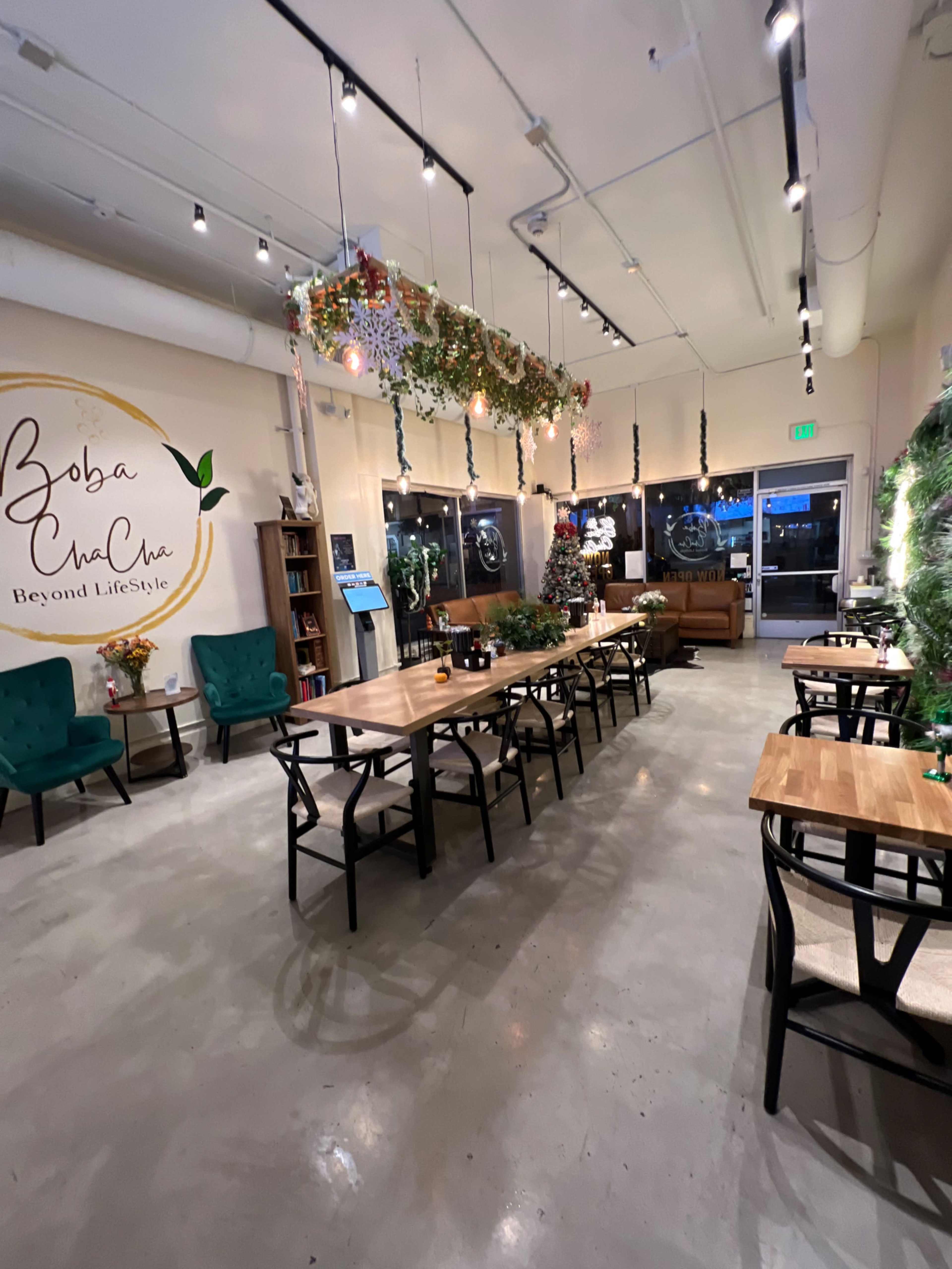 The interior of a modern café features wooden tables, green chairs, and decorative plants hanging from the ceiling, with a cozy seating area and large windows.