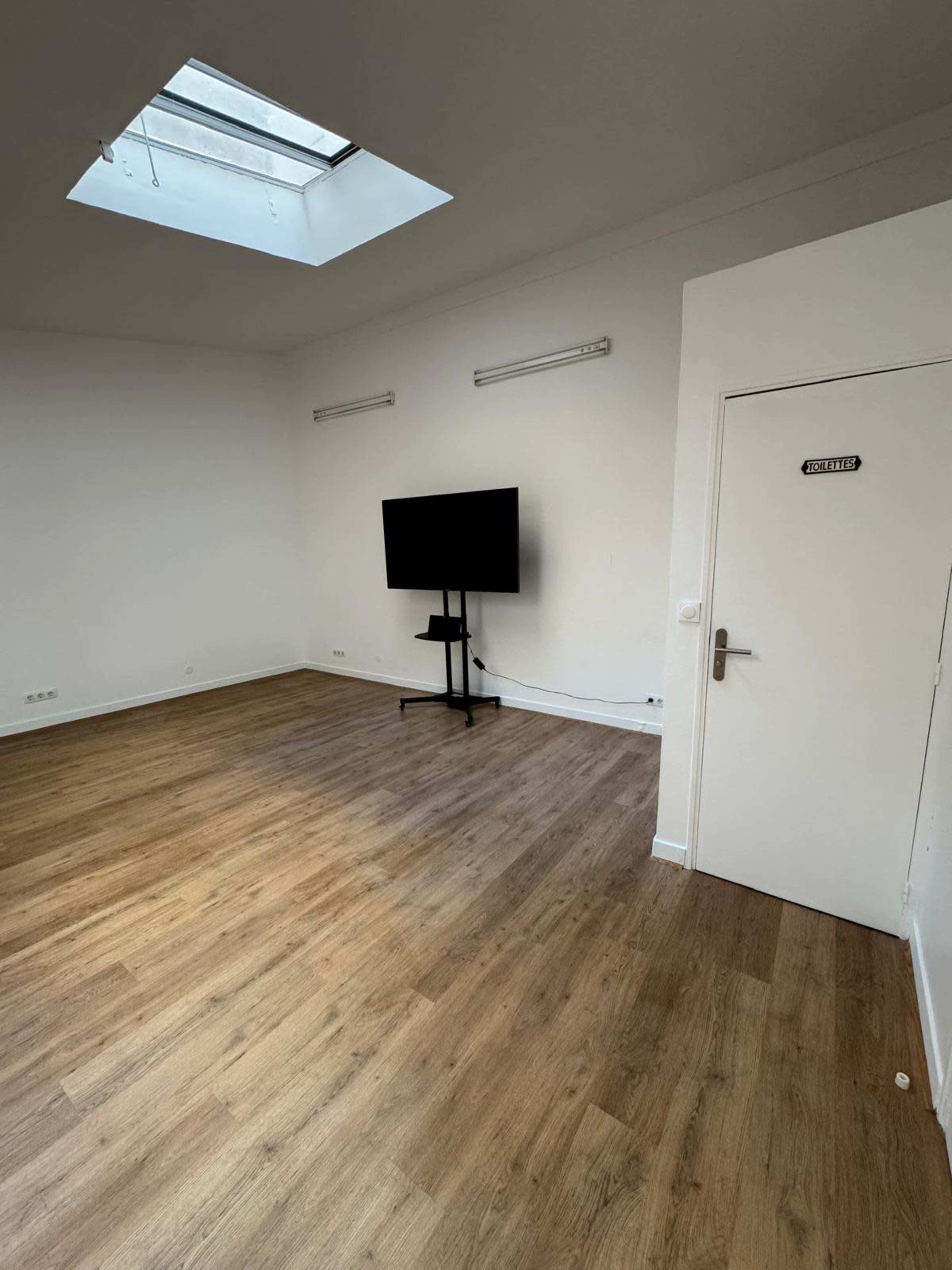 The image shows a sparse room with wooden flooring, a wall-mounted television on a stand, and a door labeled "SALLE."