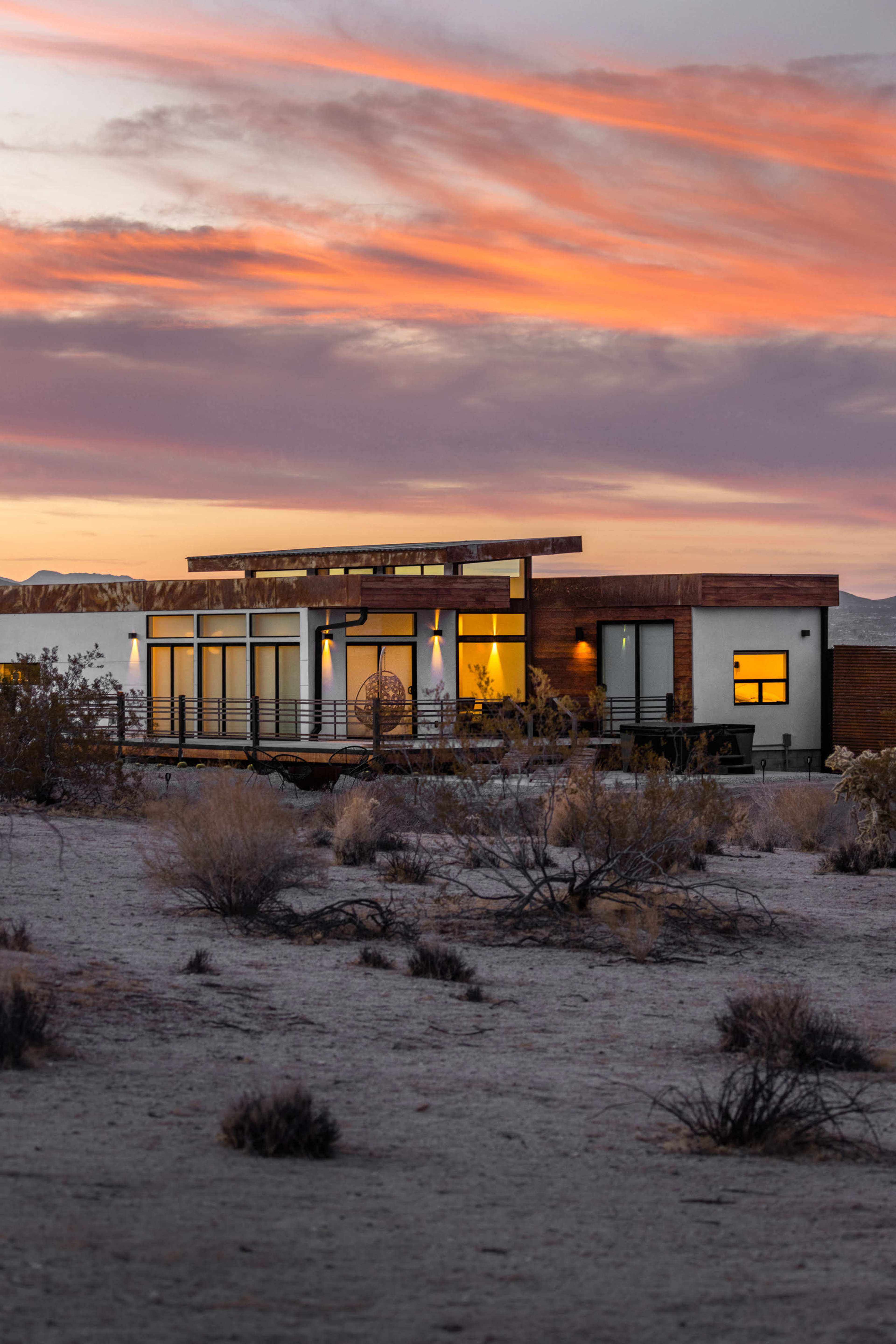 A modern house with large windows and a wooden deck is set against a colorful sunset in a desert landscape.