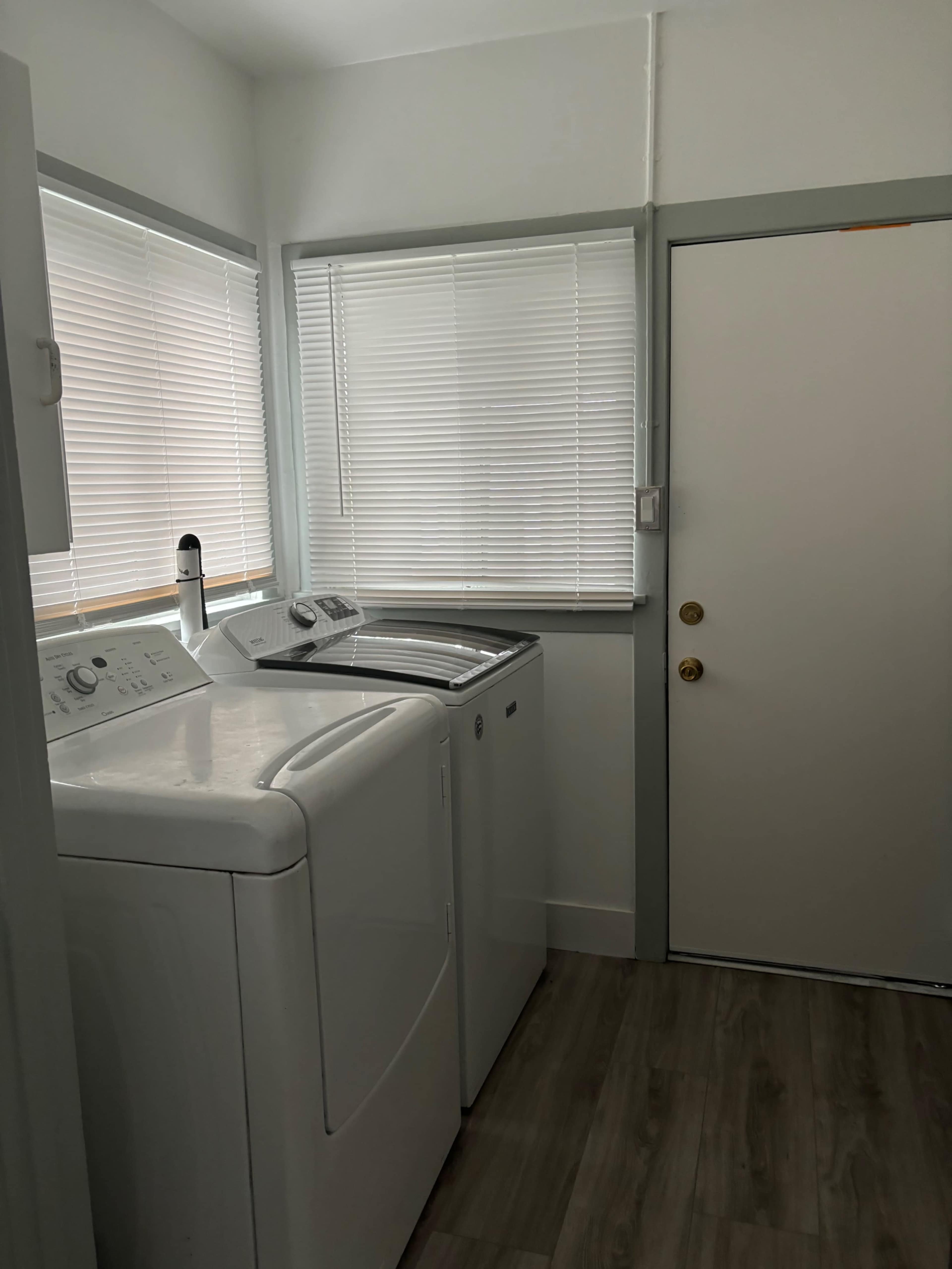 The image shows a laundry room with a washing machine and dryer beside a door, featuring white blinds on the windows.
