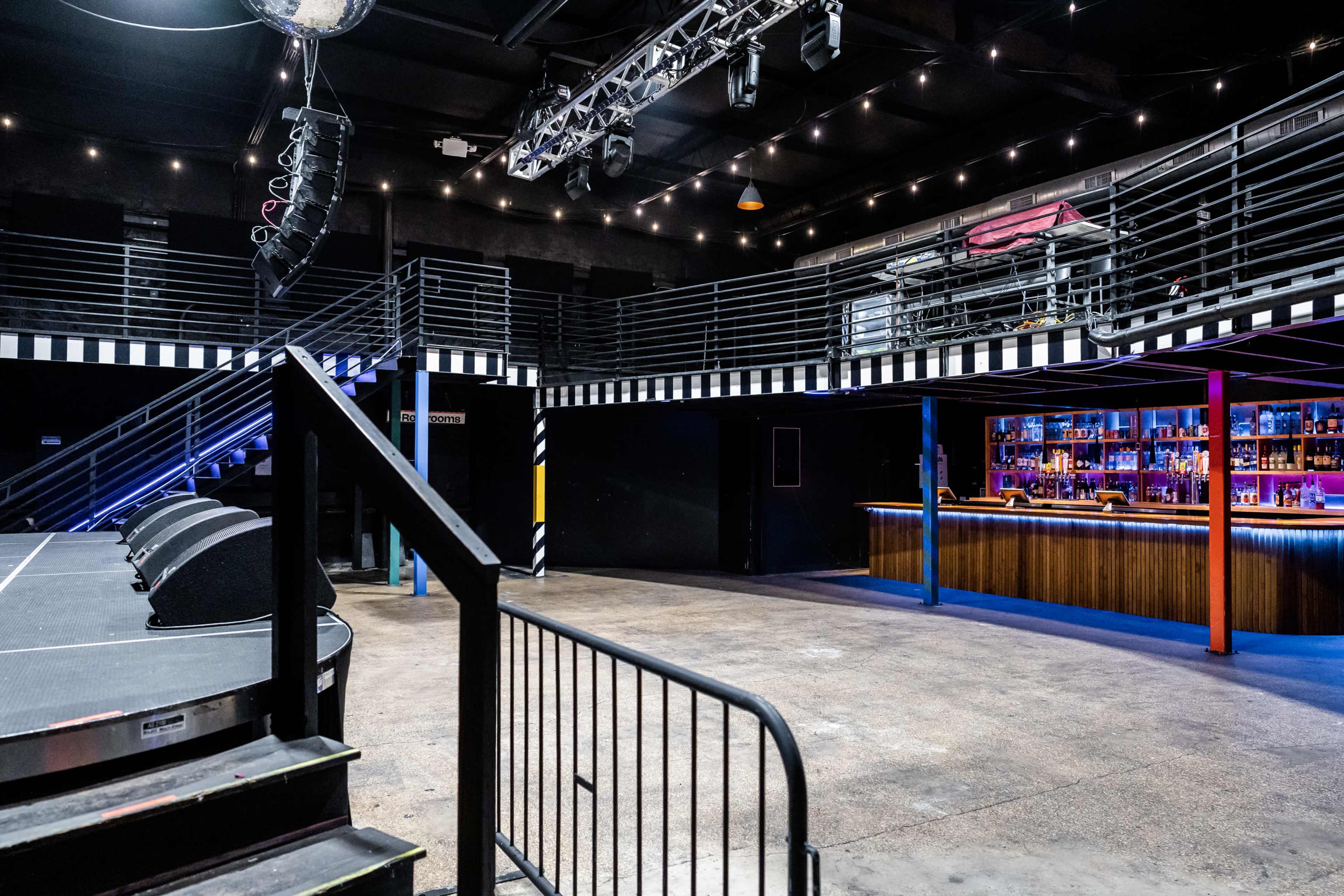 An empty music venue features a stage with speakers, a bar with illuminated shelves, and balcony seating overlooking the floor.