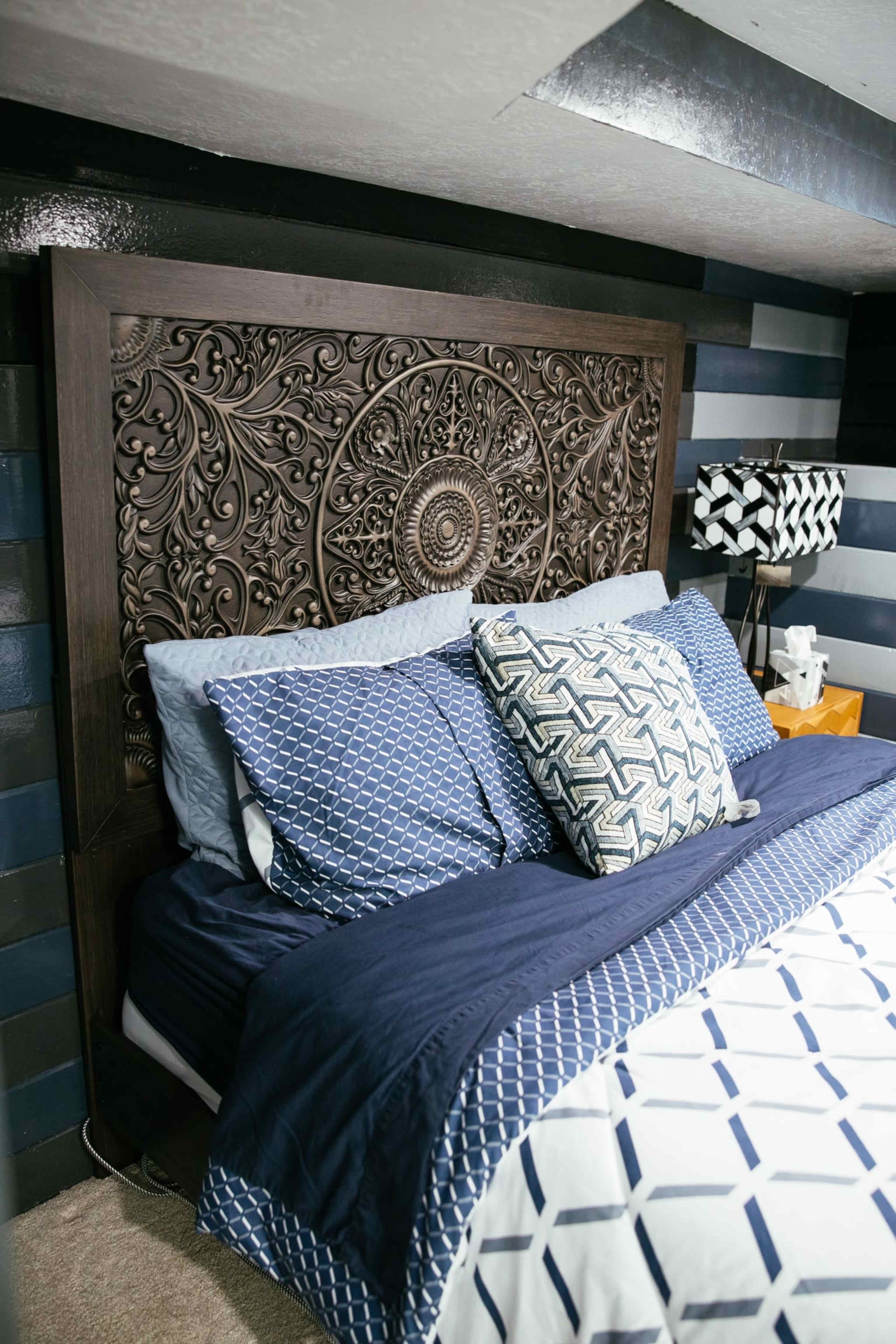 A neatly arranged bed with a carved dark wooden headboard, patterned blue and white bedding, and a nightstand with a lamp beside it.
