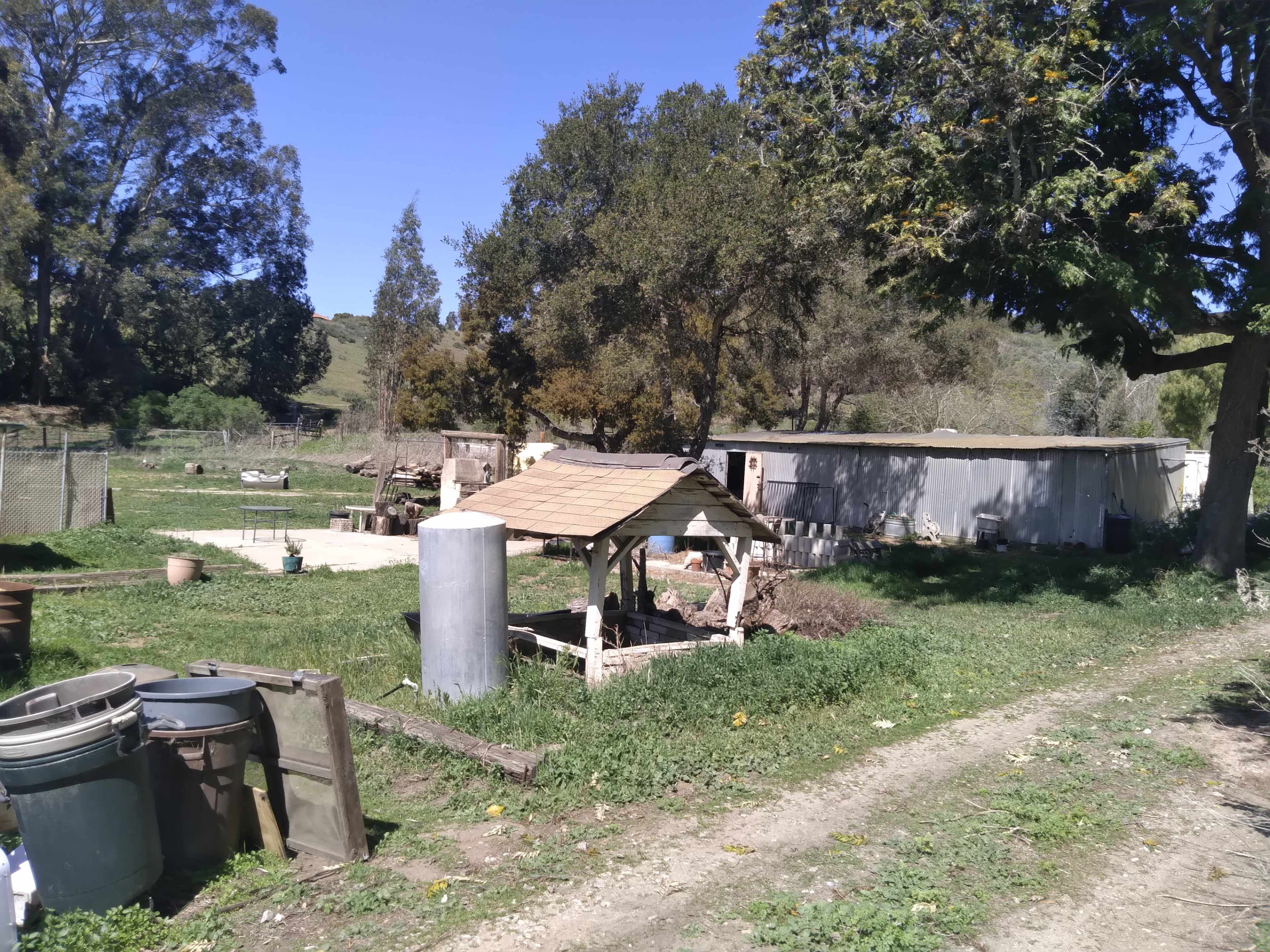A rural scene with a small wooden structure, a storage tank, and various containers on a grassy area near a mobile home.