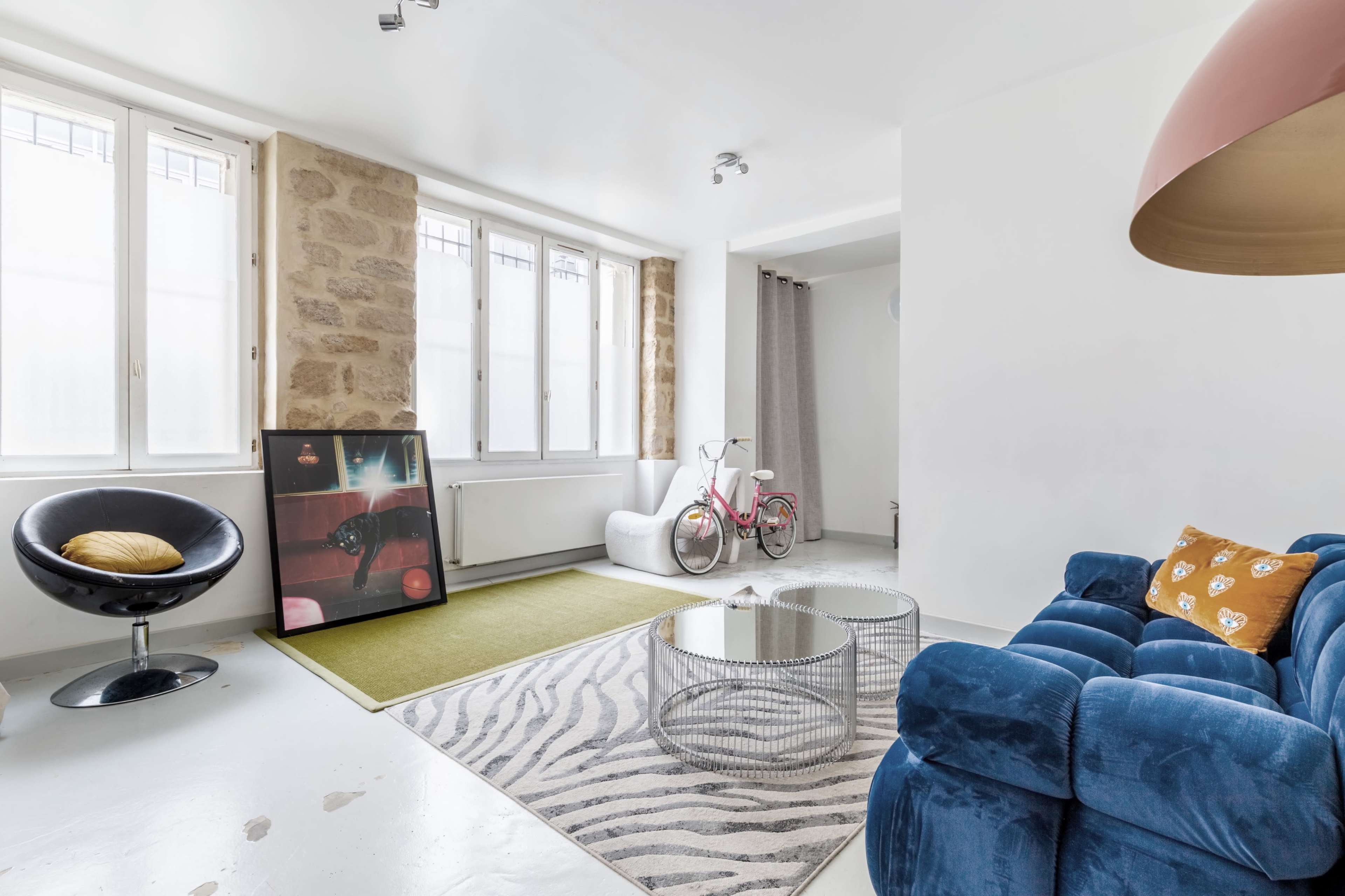 The image shows a modern living room with large windows, featuring a blue velvet sofa, a round glass coffee table, a zebra-striped rug, and a bicycle in the corner.