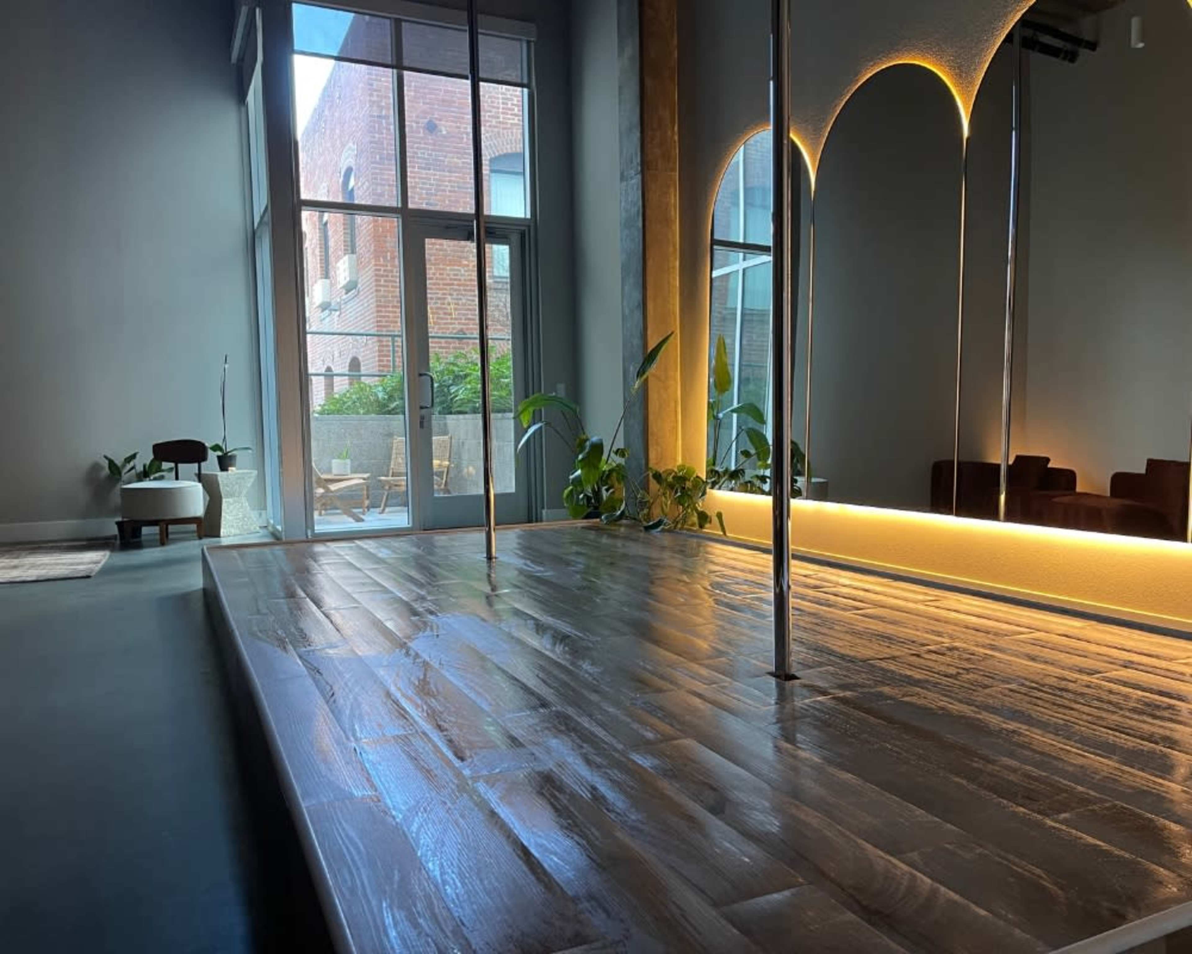 The image shows a modern interior space featuring a wooden floor, a mirrored wall, and large windows that allow natural light to enter, with plants placed around the area.