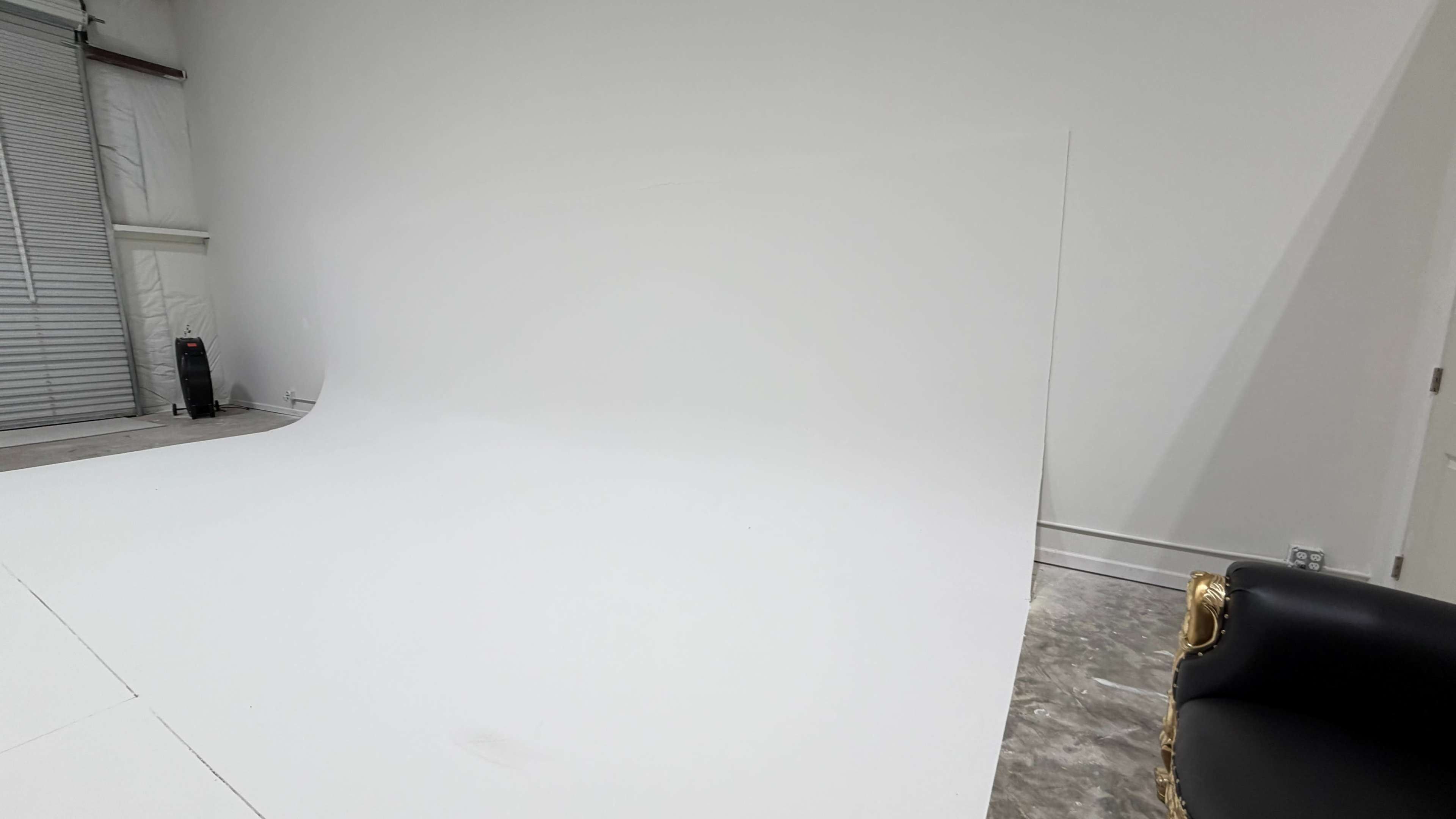 The image shows a plain, empty studio space with a large white backdrop and a corner featuring a gold chair.