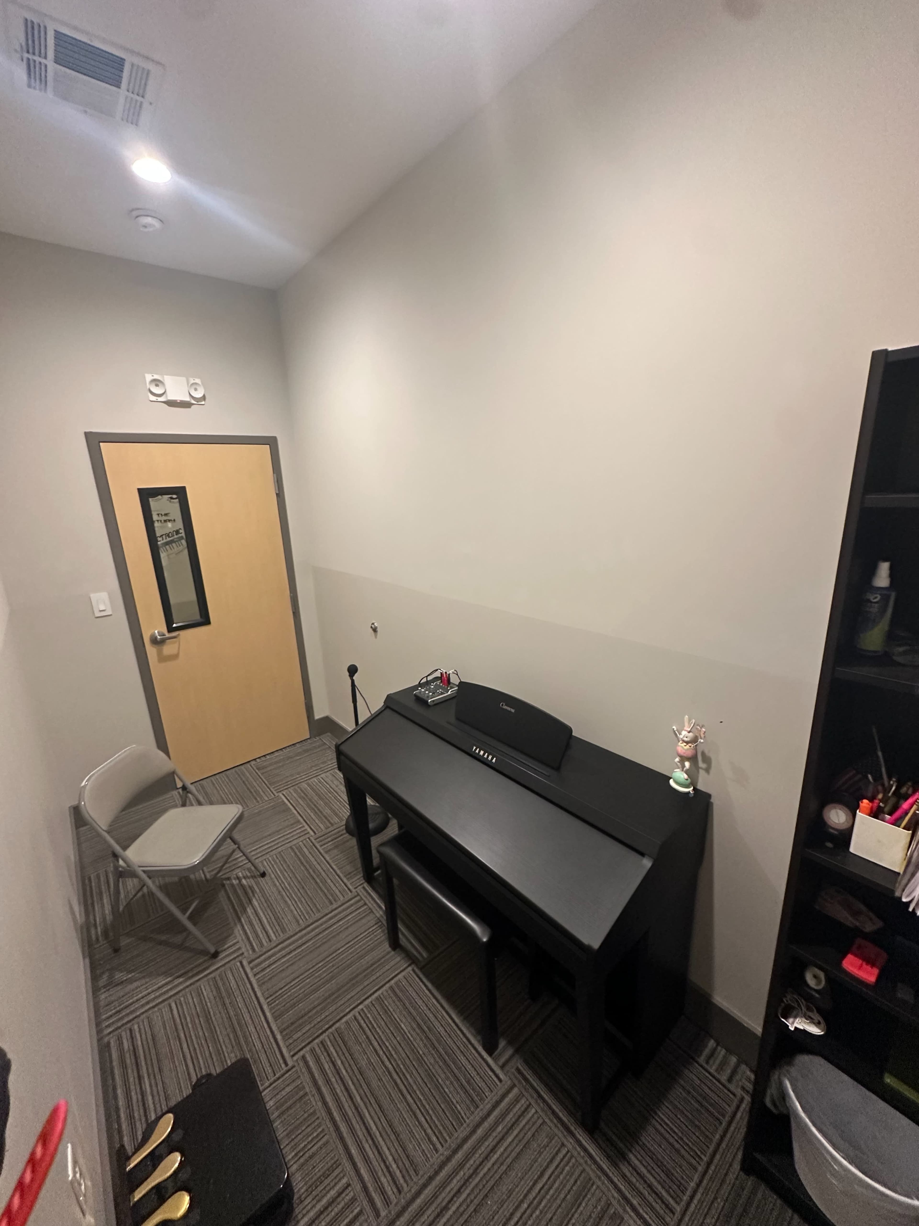 The image shows a small room featuring a black desk with a speaker, a chair, and a bookshelf filled with various items, along with a door on one side.