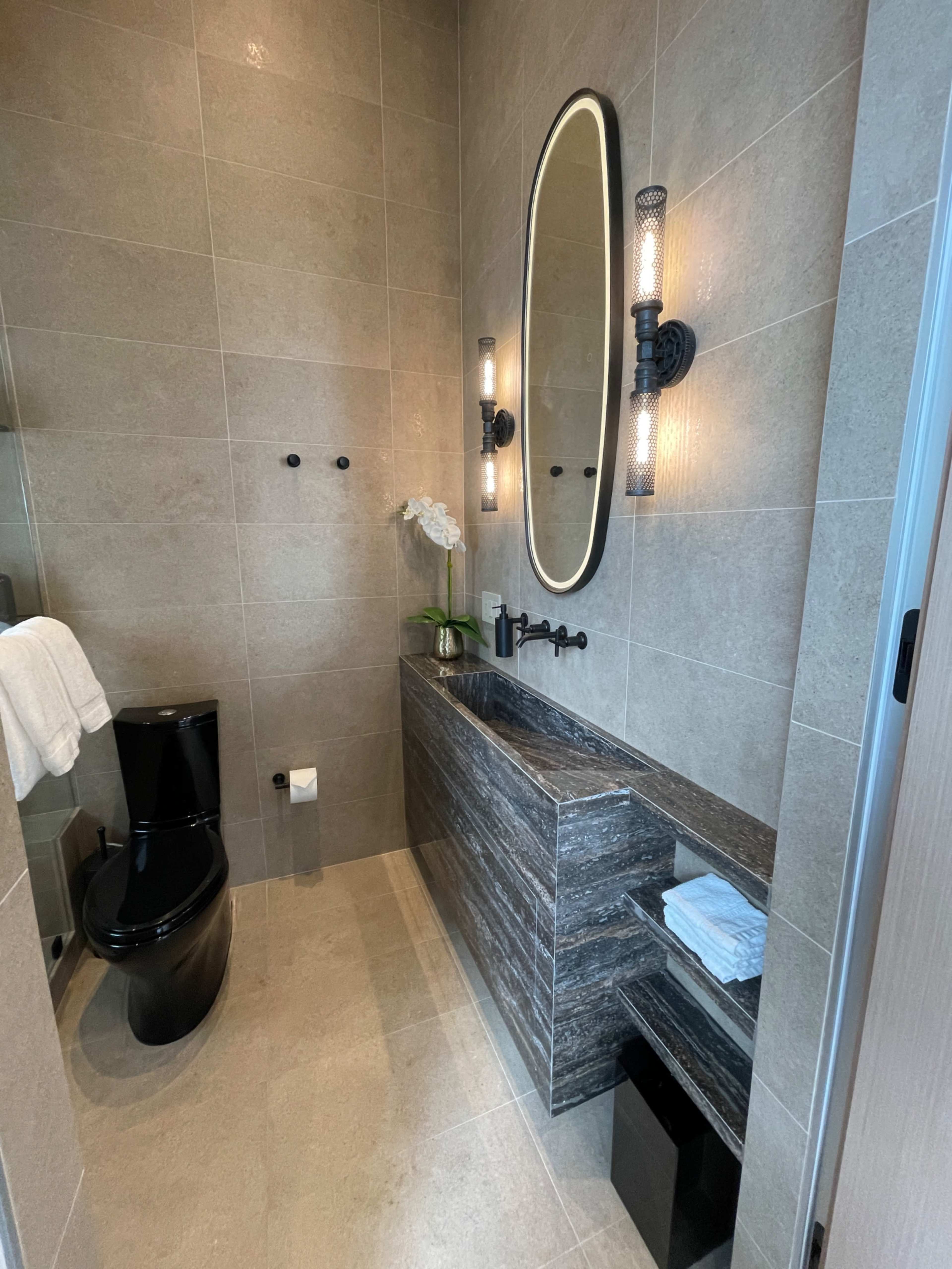 A modern bathroom featuring a sleek black toilet, a dark stone vanity with an oval mirror, and wall-mounted lights, all surrounded by muted tile walls.