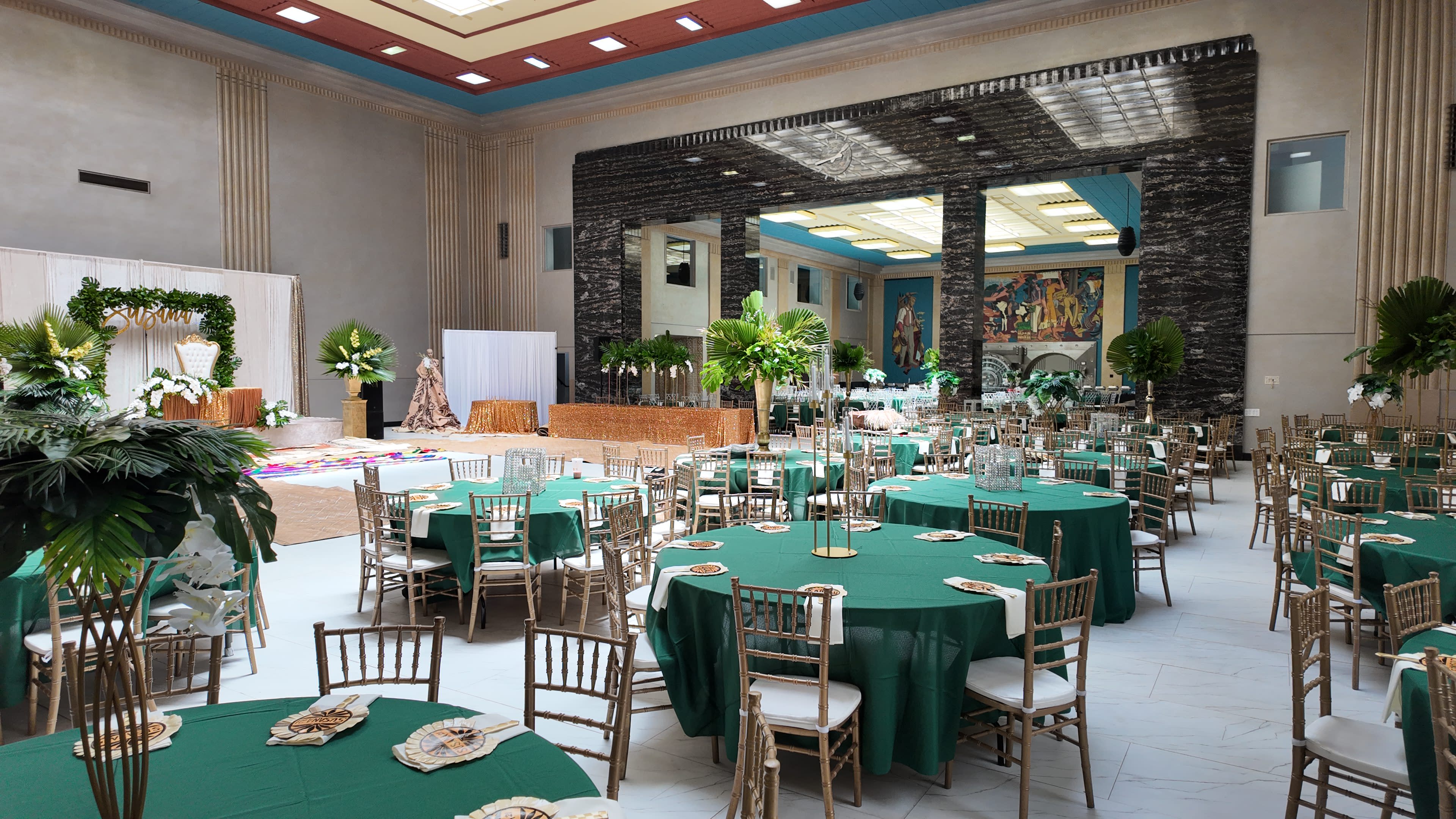 A spacious banquet hall is set up with green tablecloths and gold chairs, featuring tropical decorations and artwork on the walls.