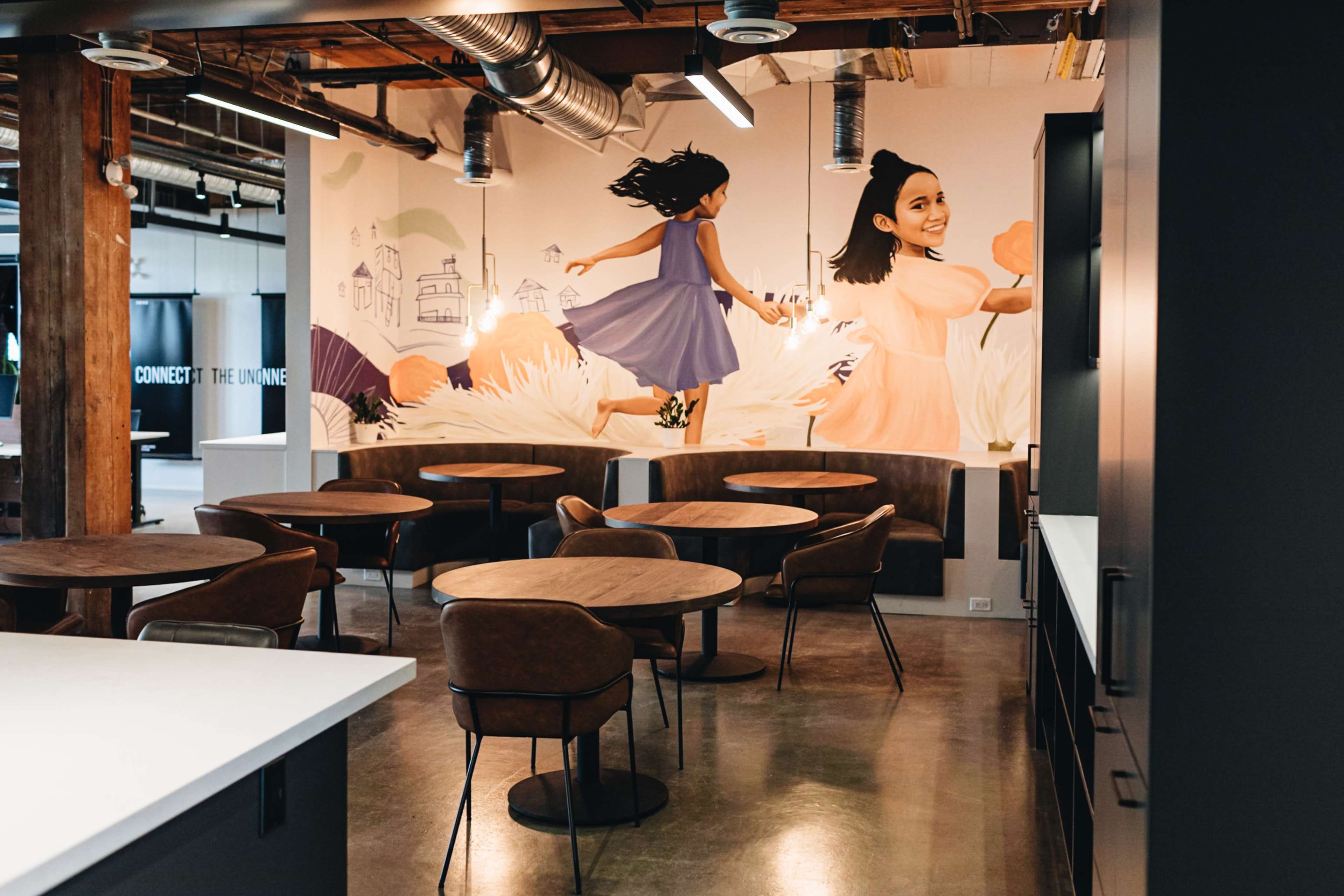 The image shows a modern office space with wooden tables and chairs, featuring a mural of a girl running on the wall behind.