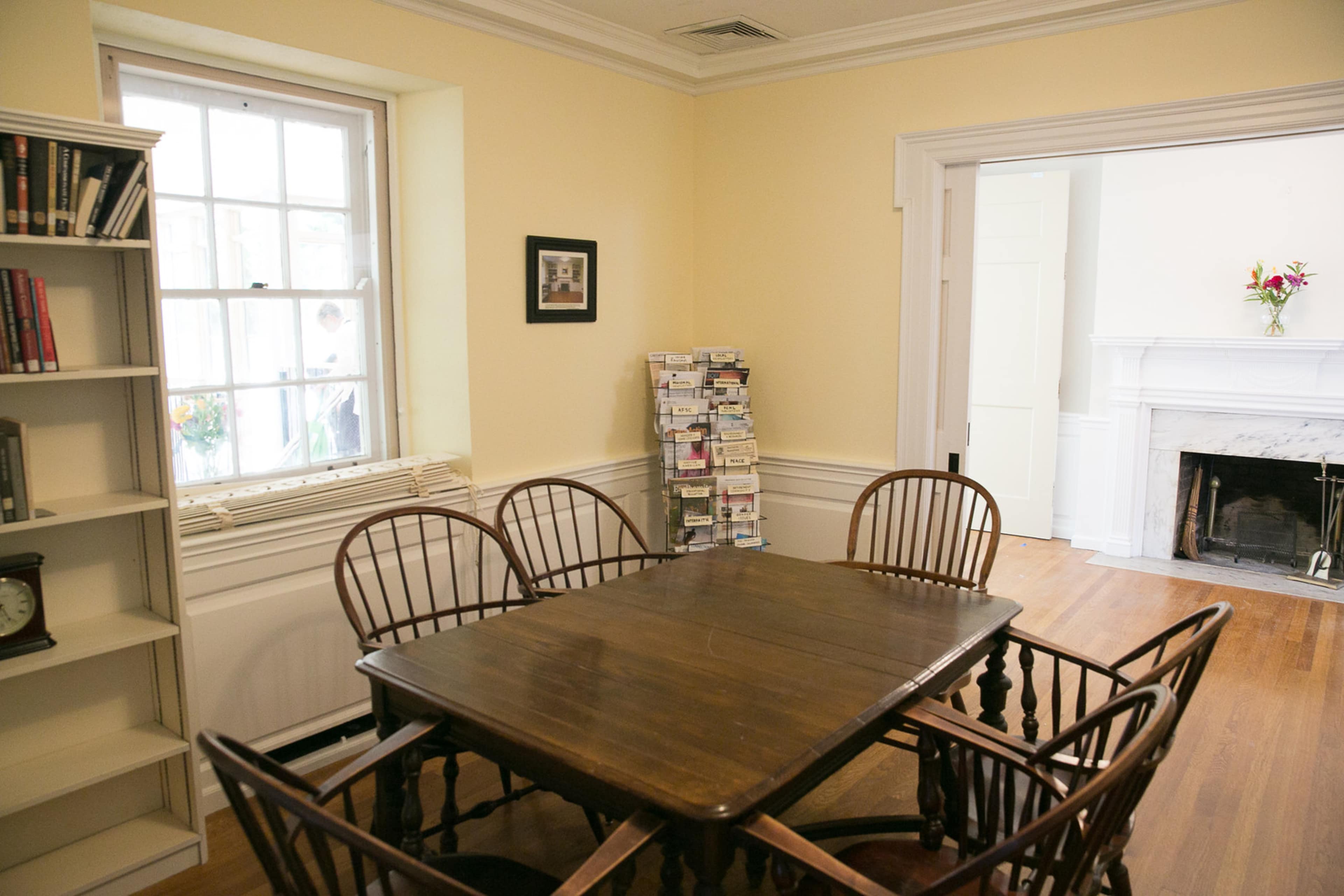 A wooden table surrounded by six chairs is positioned in a light-colored room with a window, a bookshelf, and a fireplace.