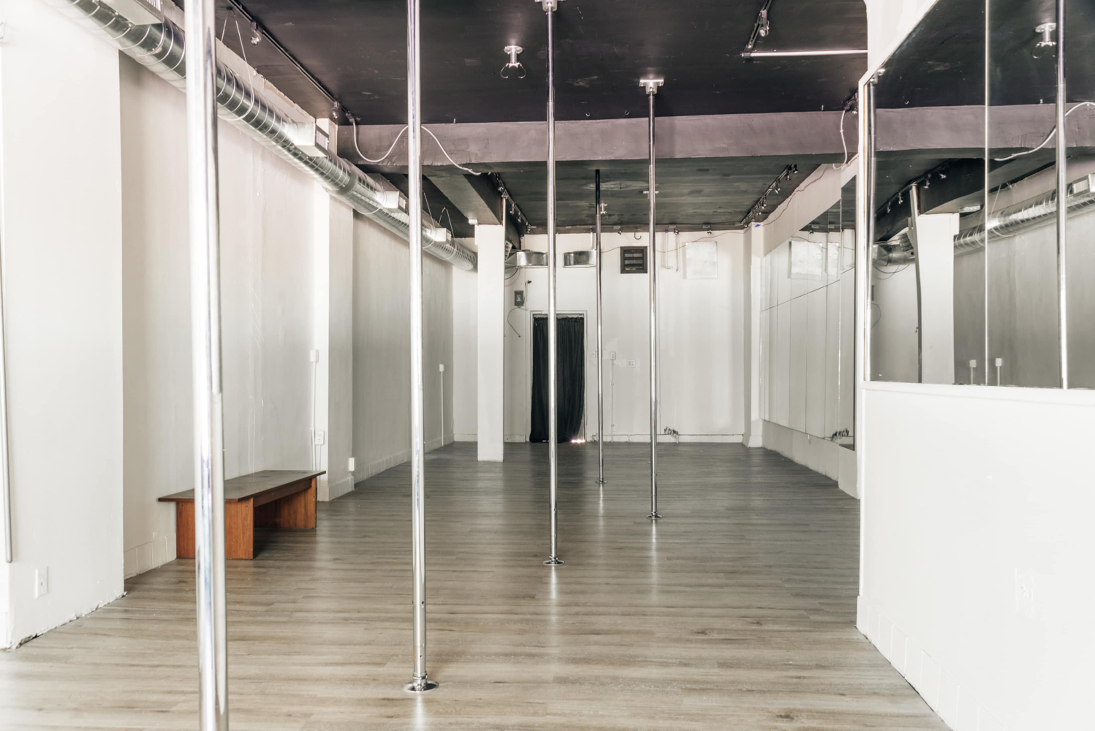 The interior of a spacious dance studio features several vertical poles and mirrors along the walls, with wooden seating in one corner.