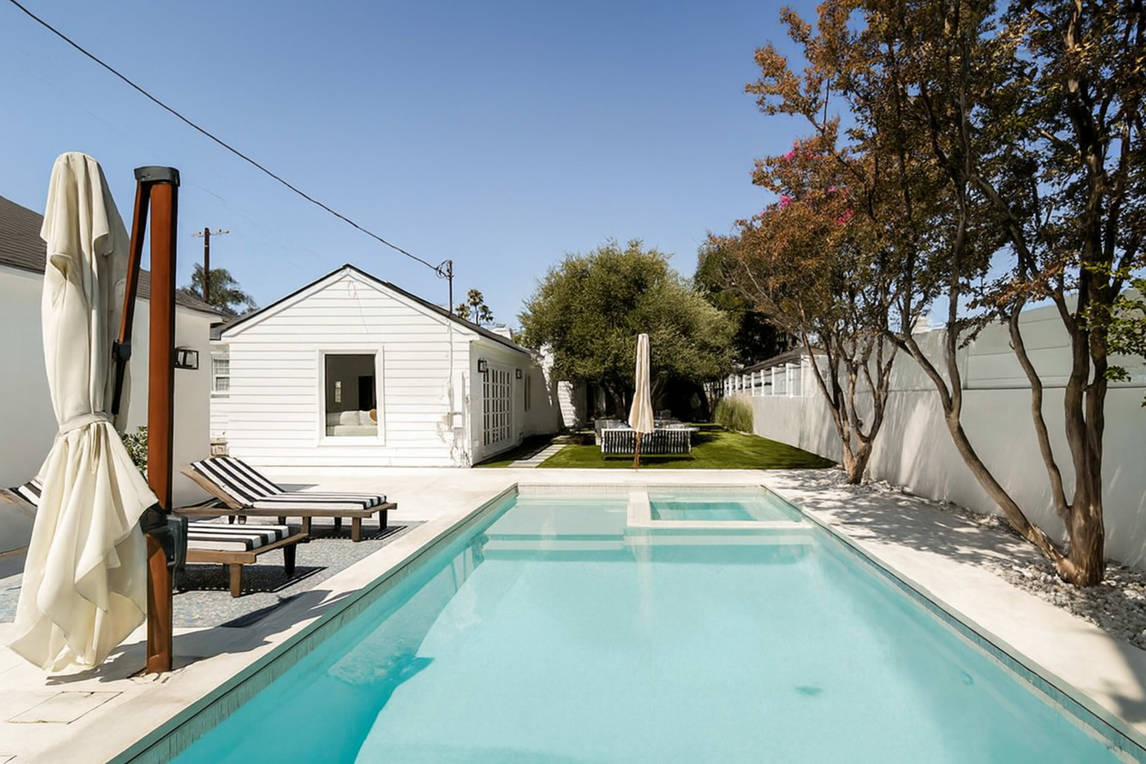A clear blue pool is situated in a backyard with lounge chairs and a small white building in the background, framed by green trees and grass.