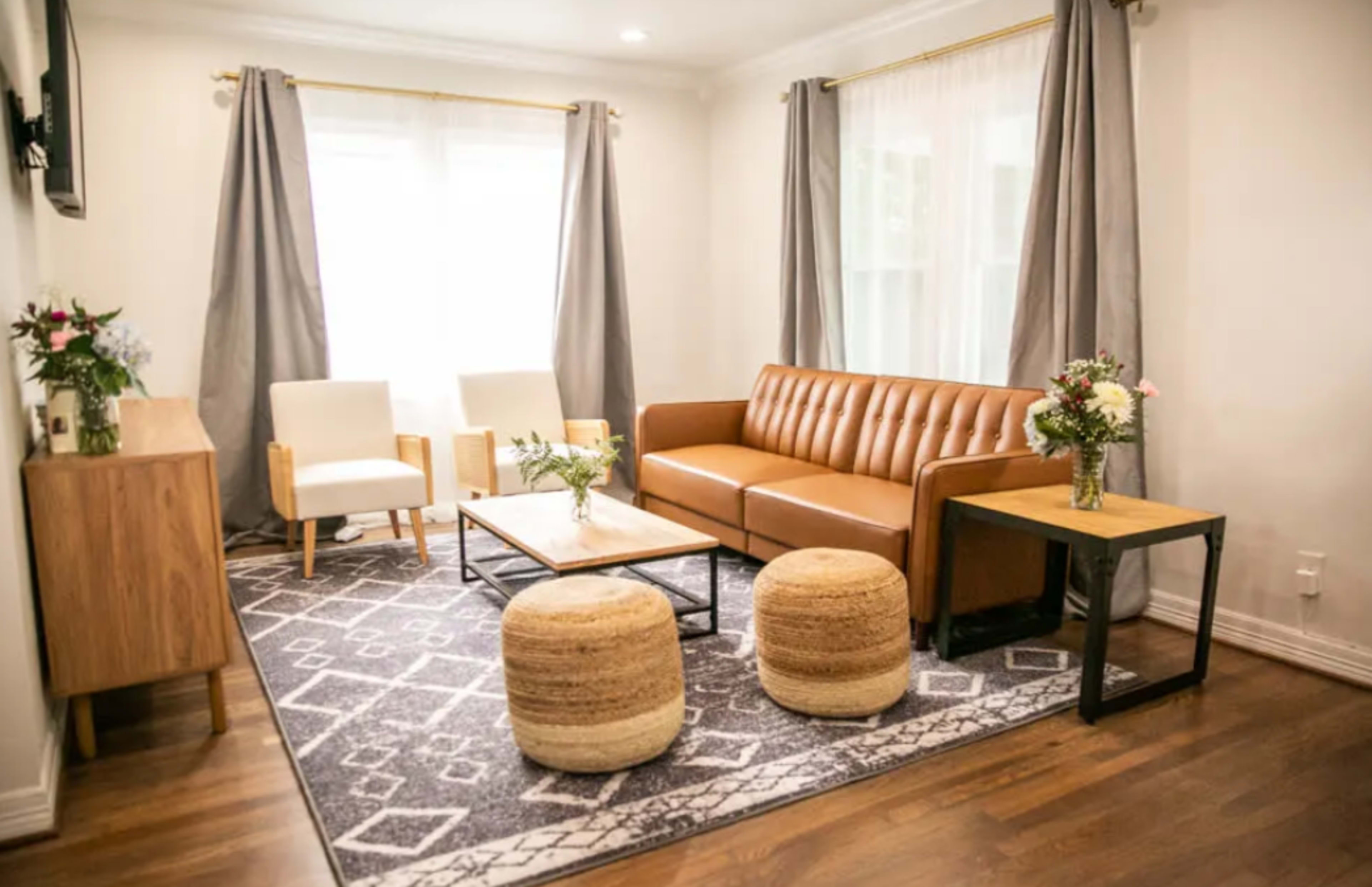 A modern living room with a brown leather sofa, two white chairs, a coffee table, and a woven area rug, surrounded by large windows with light gray curtains.