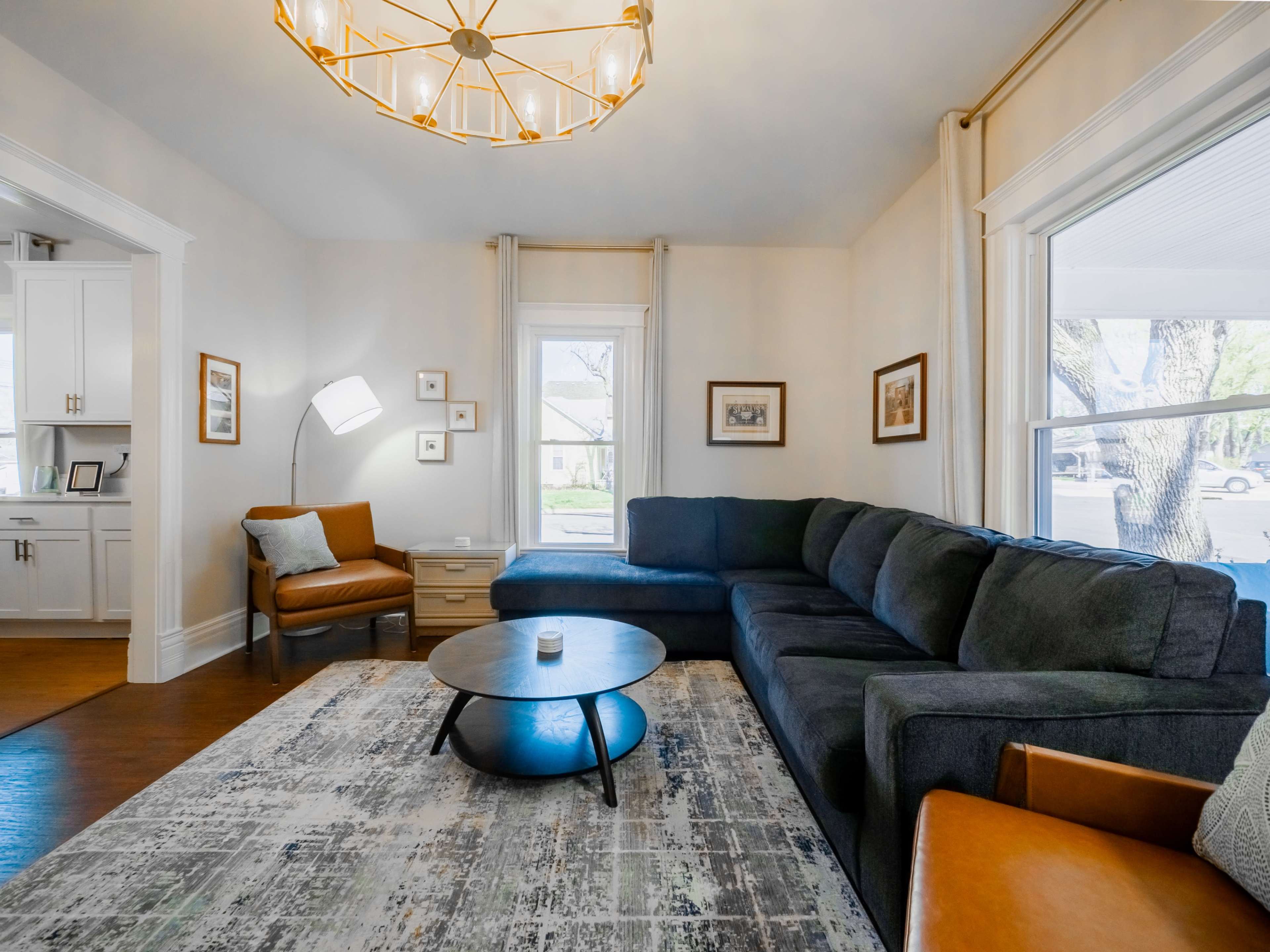 The living room features a large sectional sofa, a round coffee table, and a modern light fixture, with windows allowing natural light to enter.