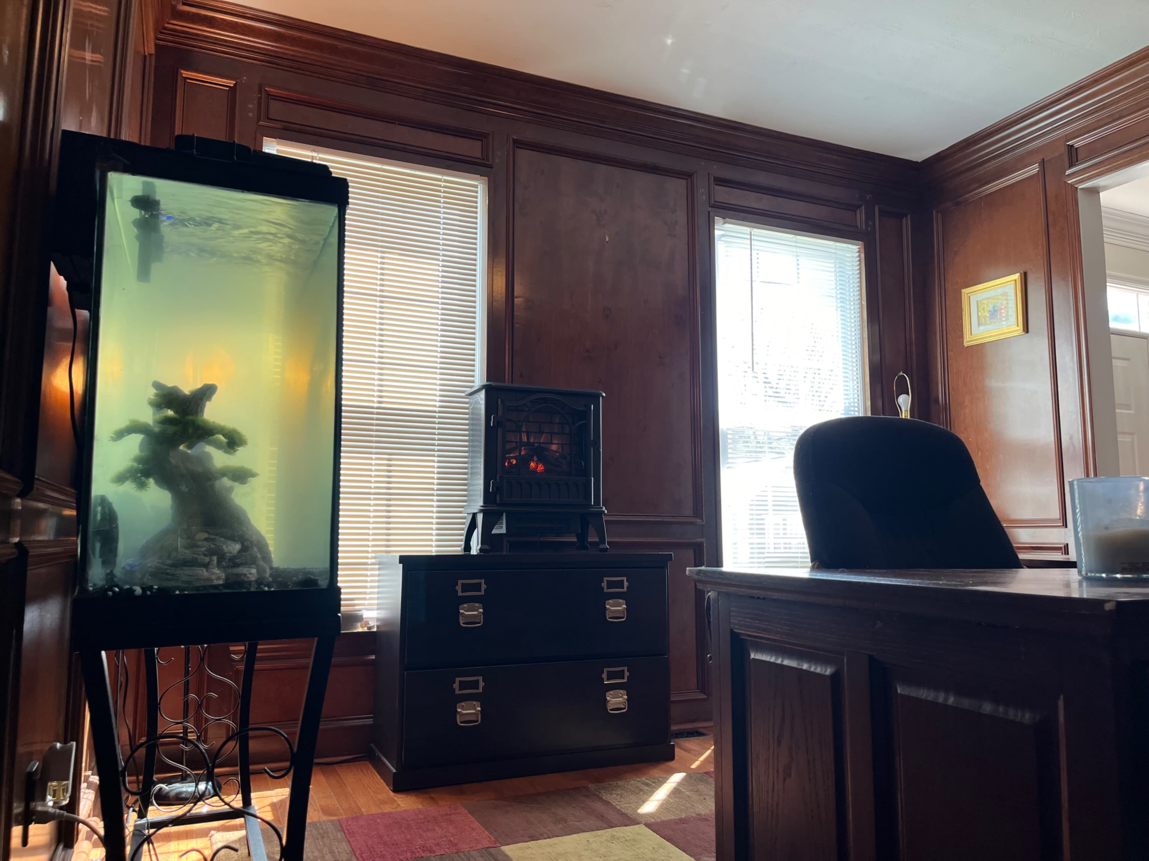 A well-furnished office with a fish tank, a black heater, a wooden desk, and large windows allowing natural light to enter.