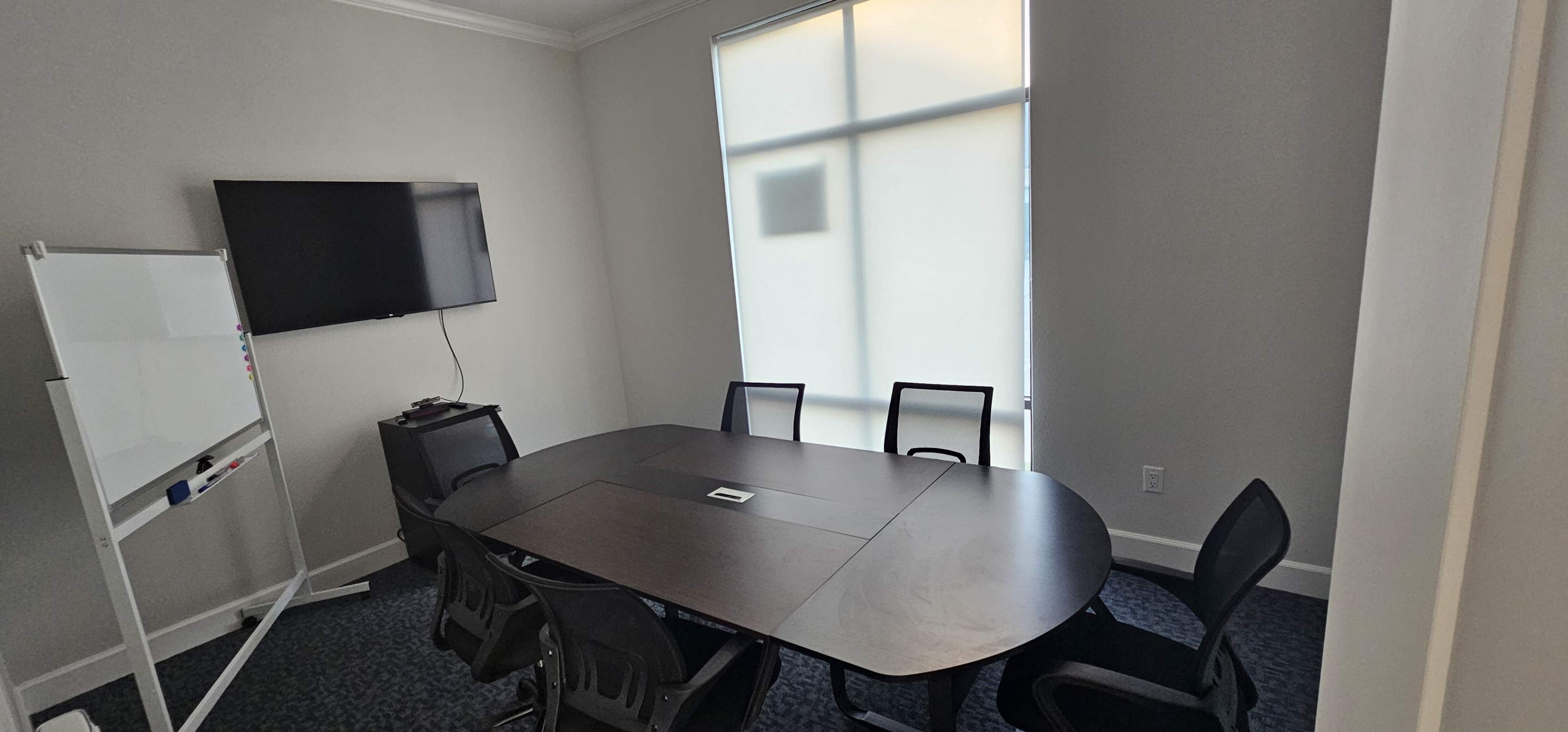 A modern conference room features a large table surrounded by six chairs, a wall-mounted television, and a whiteboard.