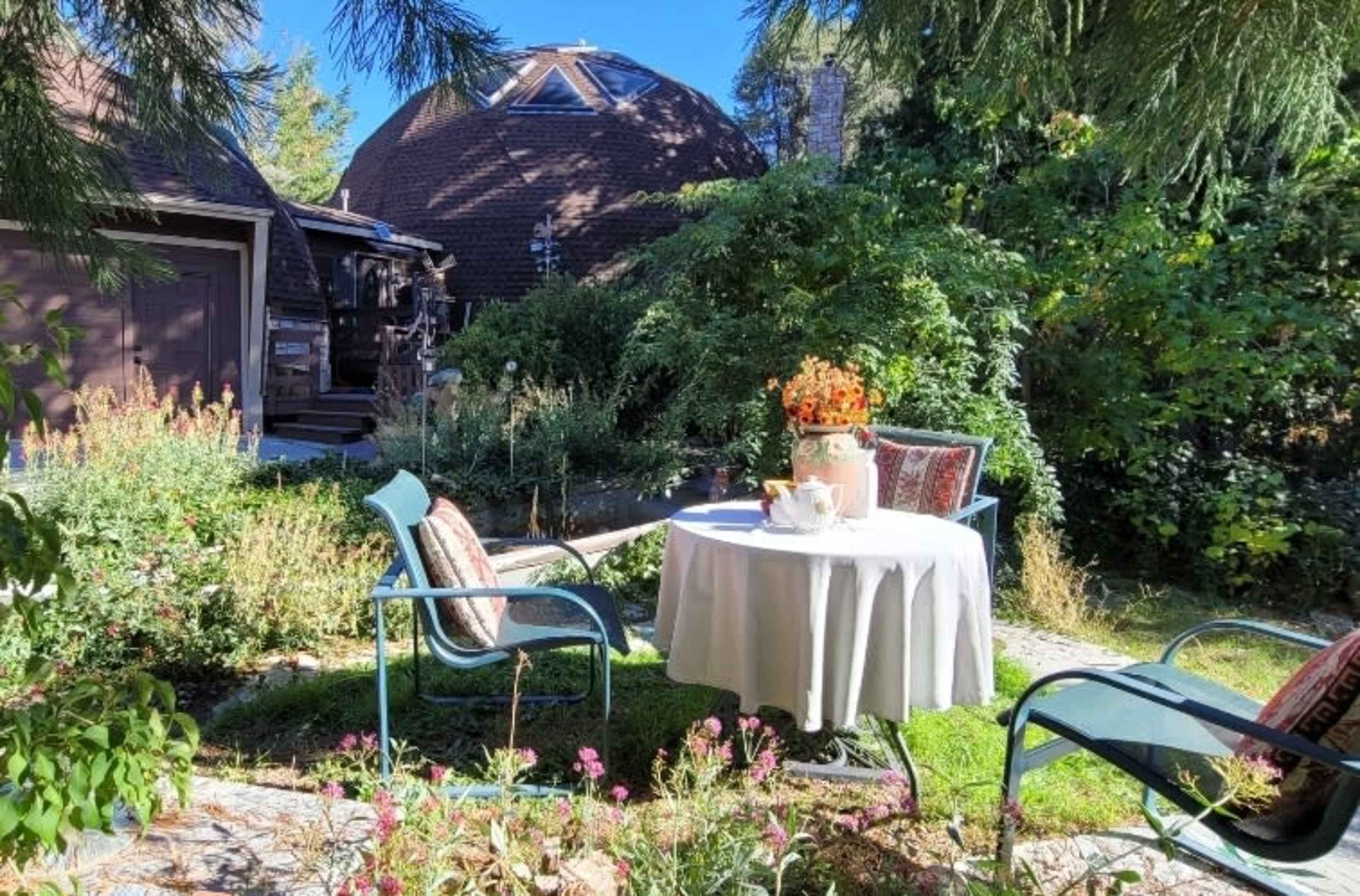 A small outdoor seating area features a round table with a tablecloth, surrounded by two chairs, set amongst lush greenery and flowers.