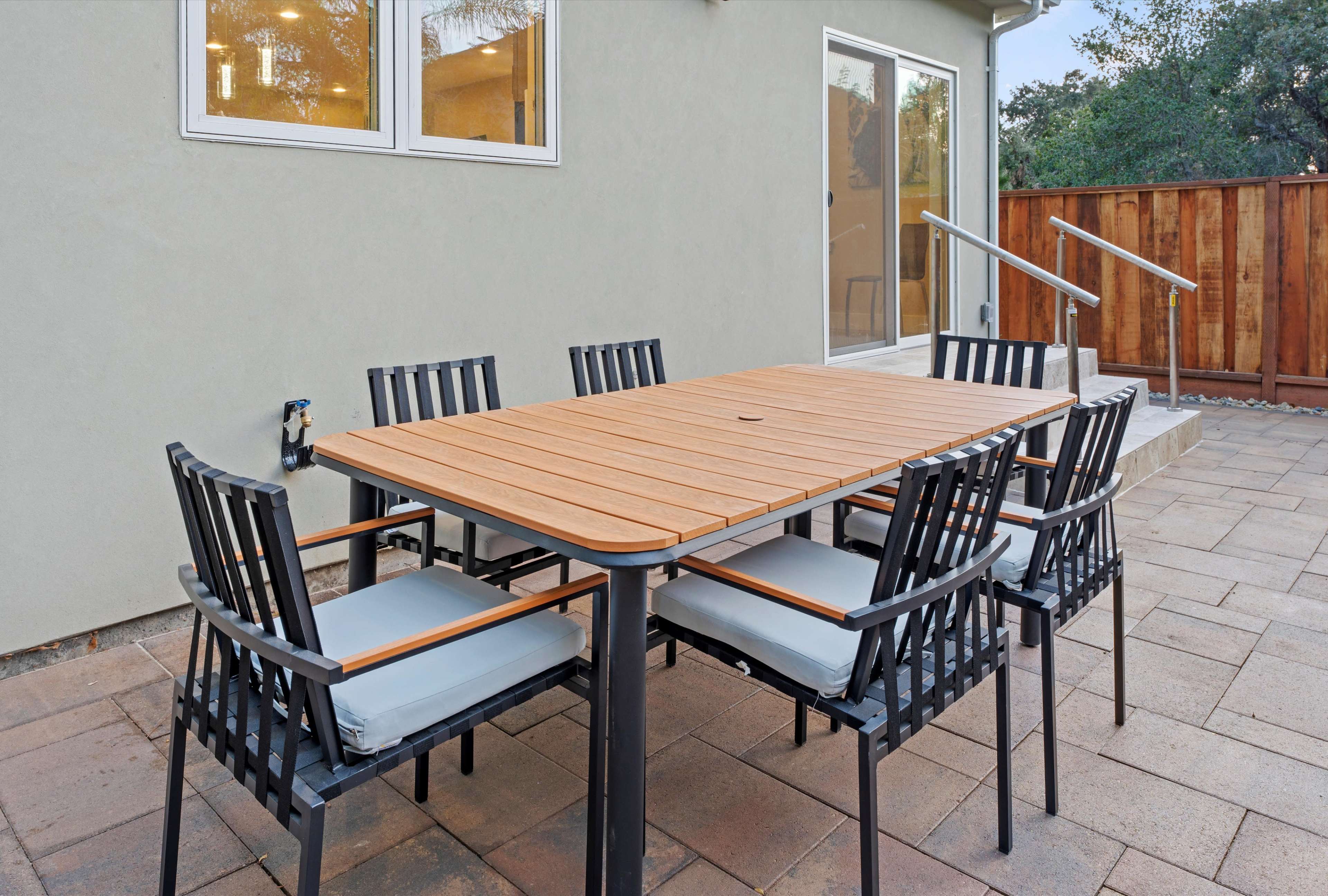 A rectangular wooden table with six black chairs is set on a stone patio beside a house with a sliding glass door.