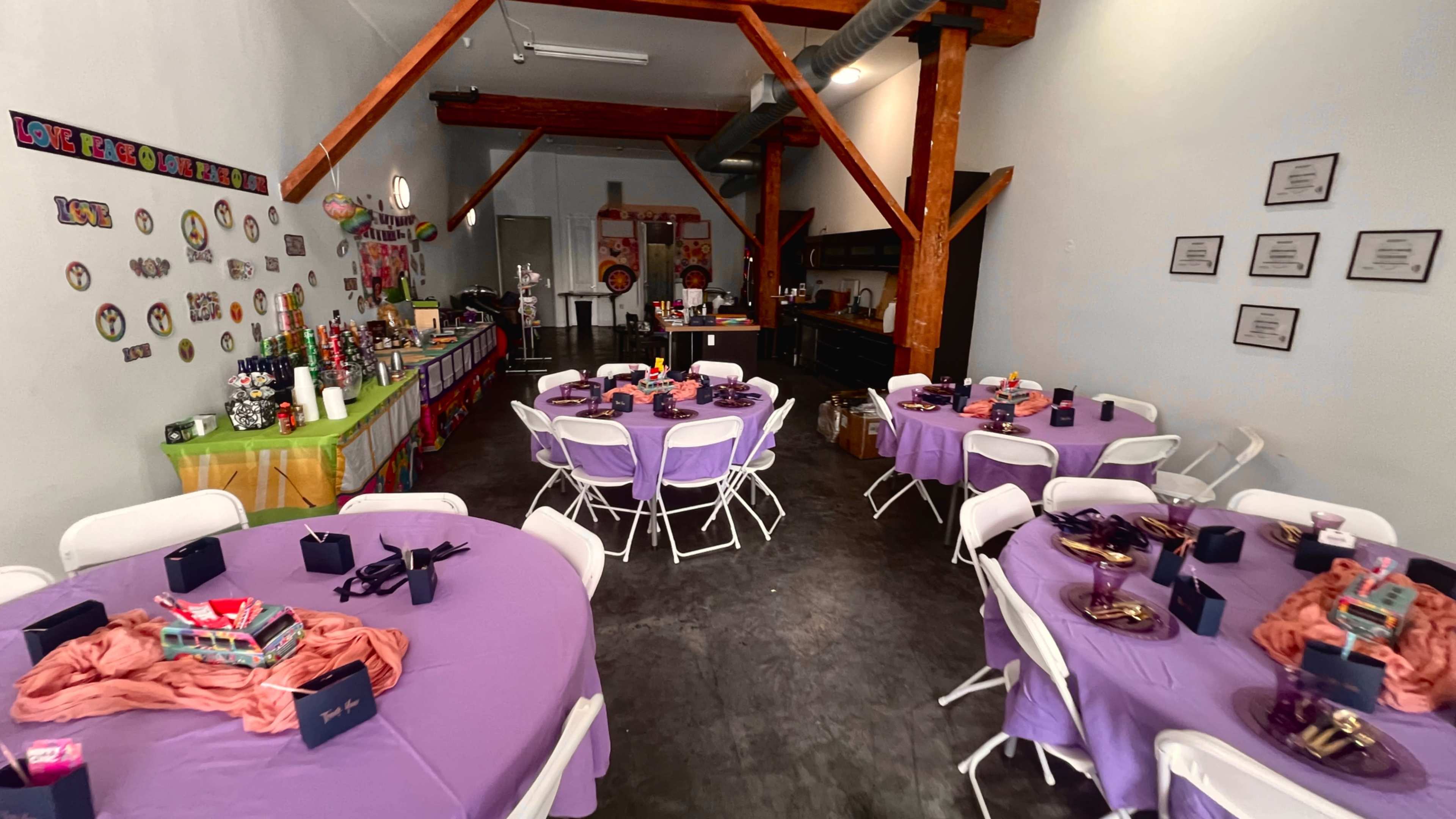 The image shows a decorated event space with several round tables covered in purple tablecloths, set with party favors and surrounded by colorful wall decorations.