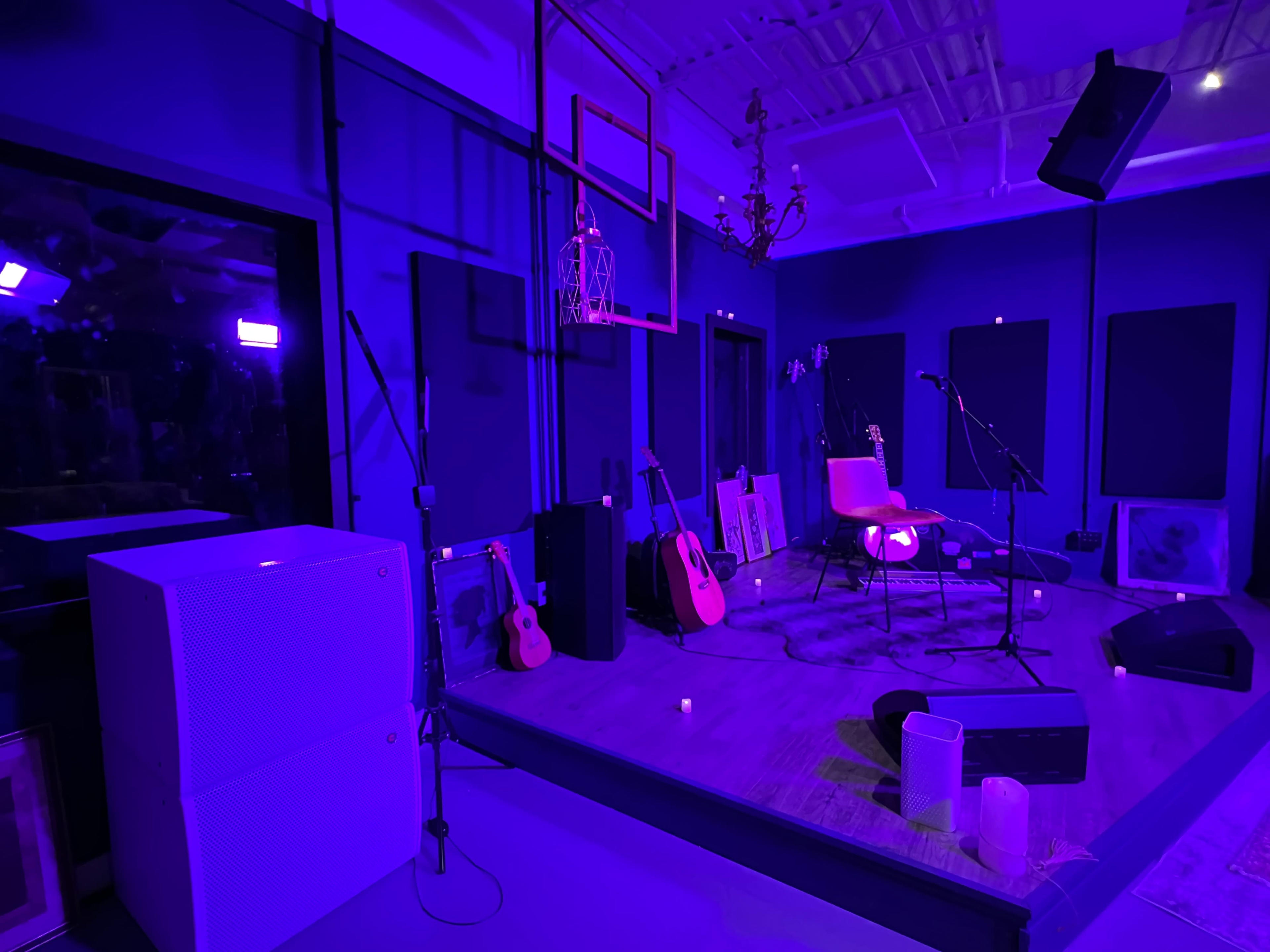 A dimly lit stage setup with musical instruments, including guitars, microphones, and a microphone stand, surrounded by purple lighting.