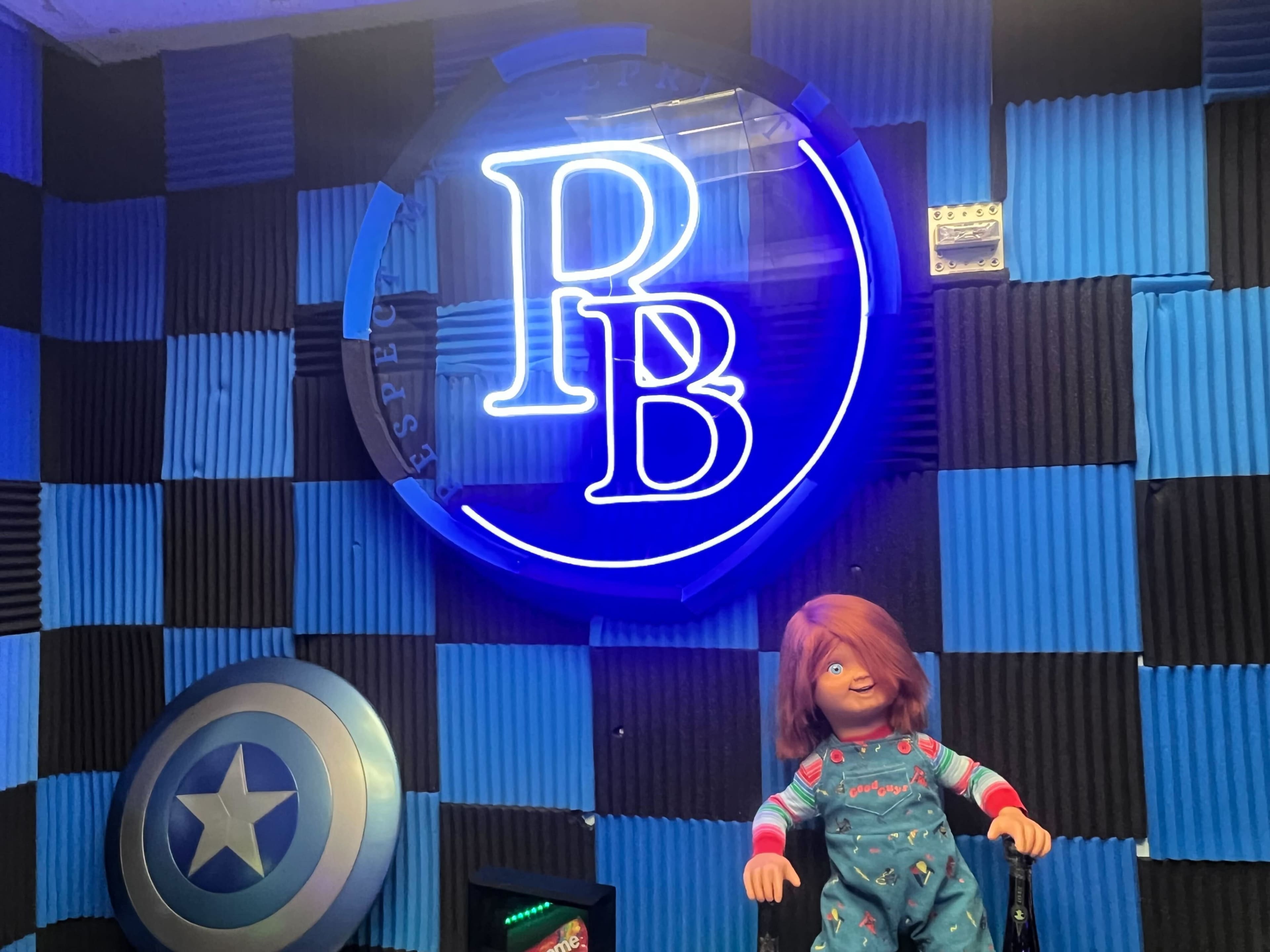 A brightly lit room with a neon sign displaying the letters "PB" and a doll with red hair sitting in front of a shield-shaped decoration.