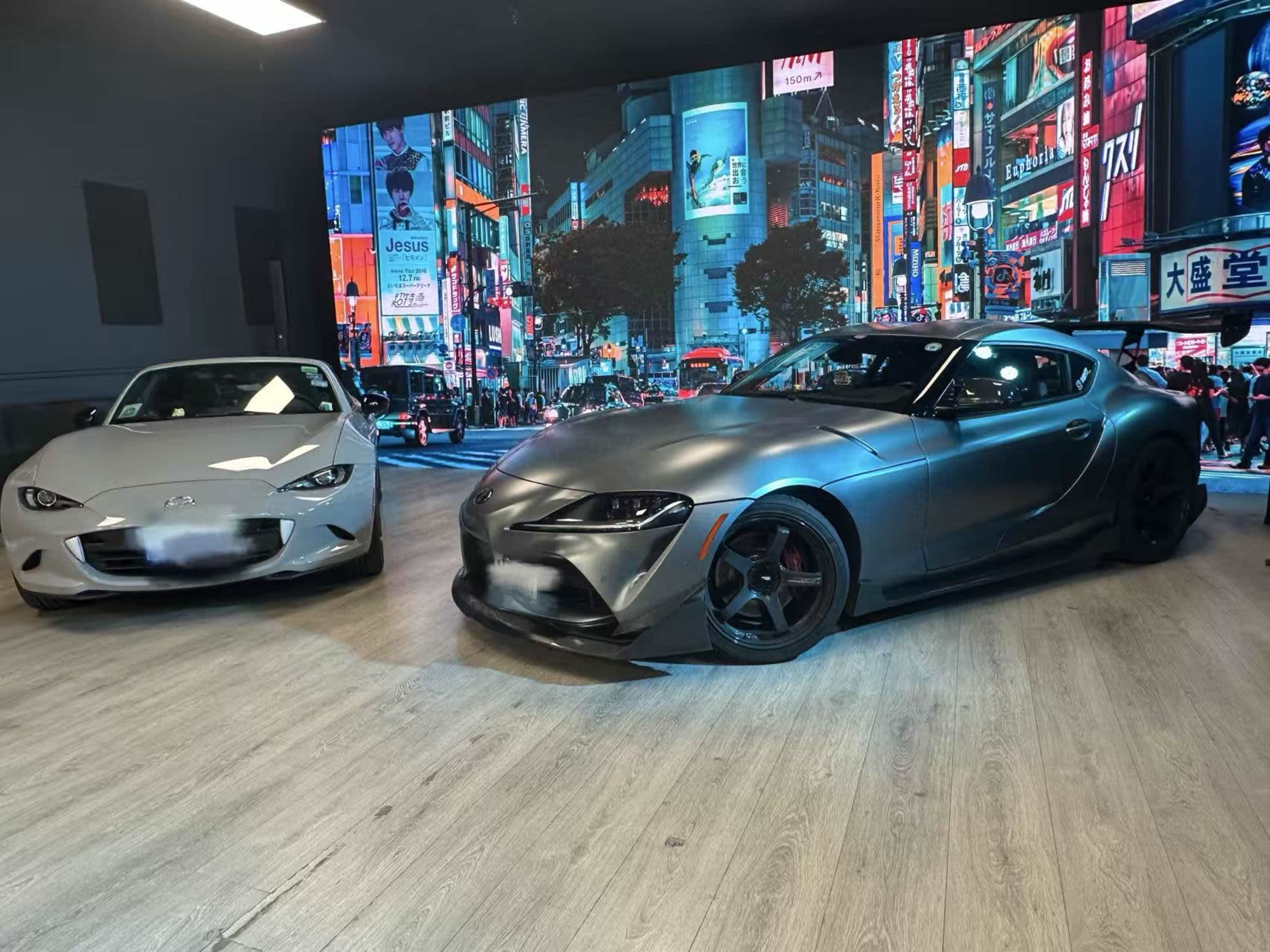 A gray sports car is parked next to a matte black sports car in a showroom featuring a vibrant cityscape backdrop.