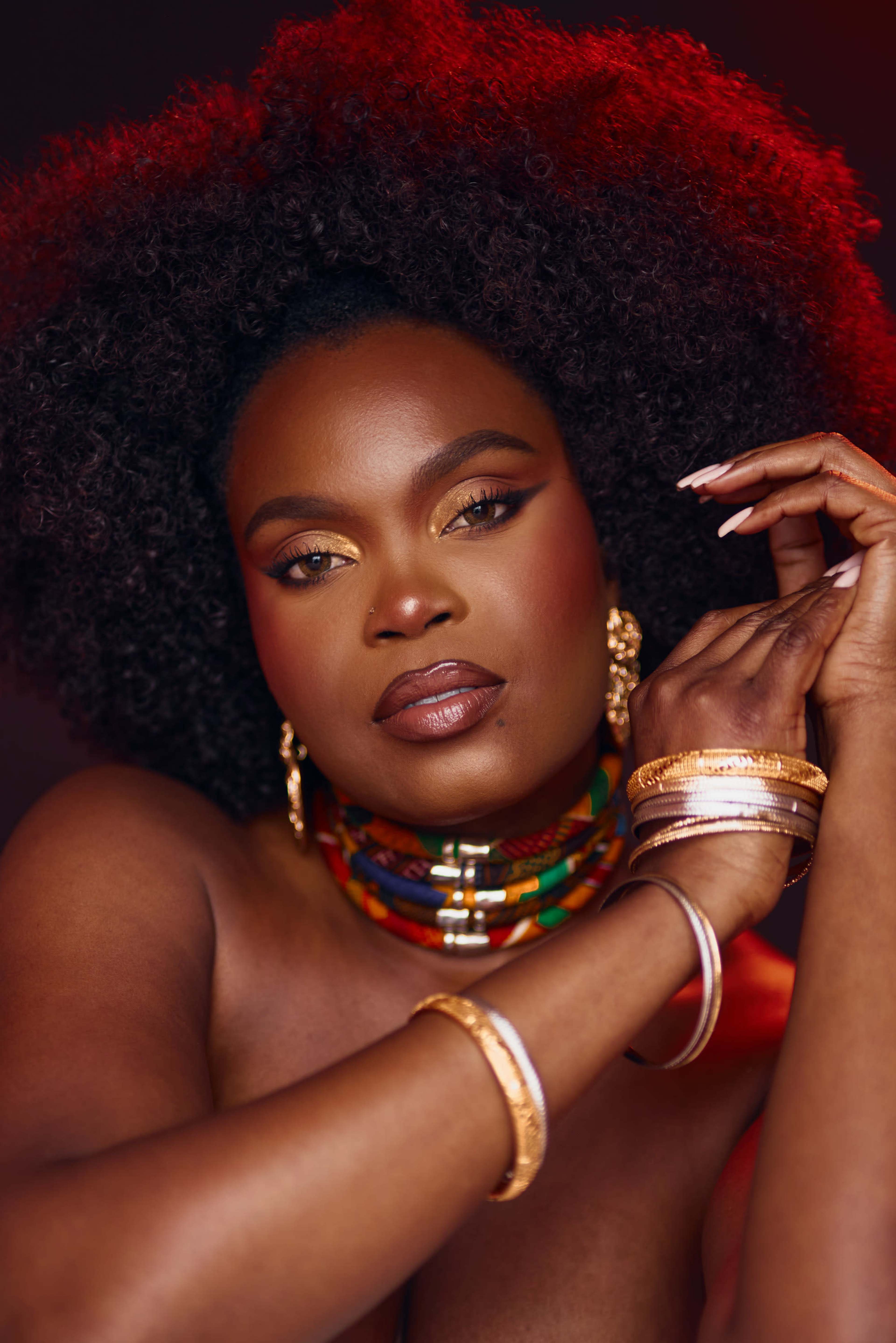 A woman with a voluminous afro hairstyle poses elegantly, adorned with colorful jewelry and gold bracelets, against a dark, softly lit background.
