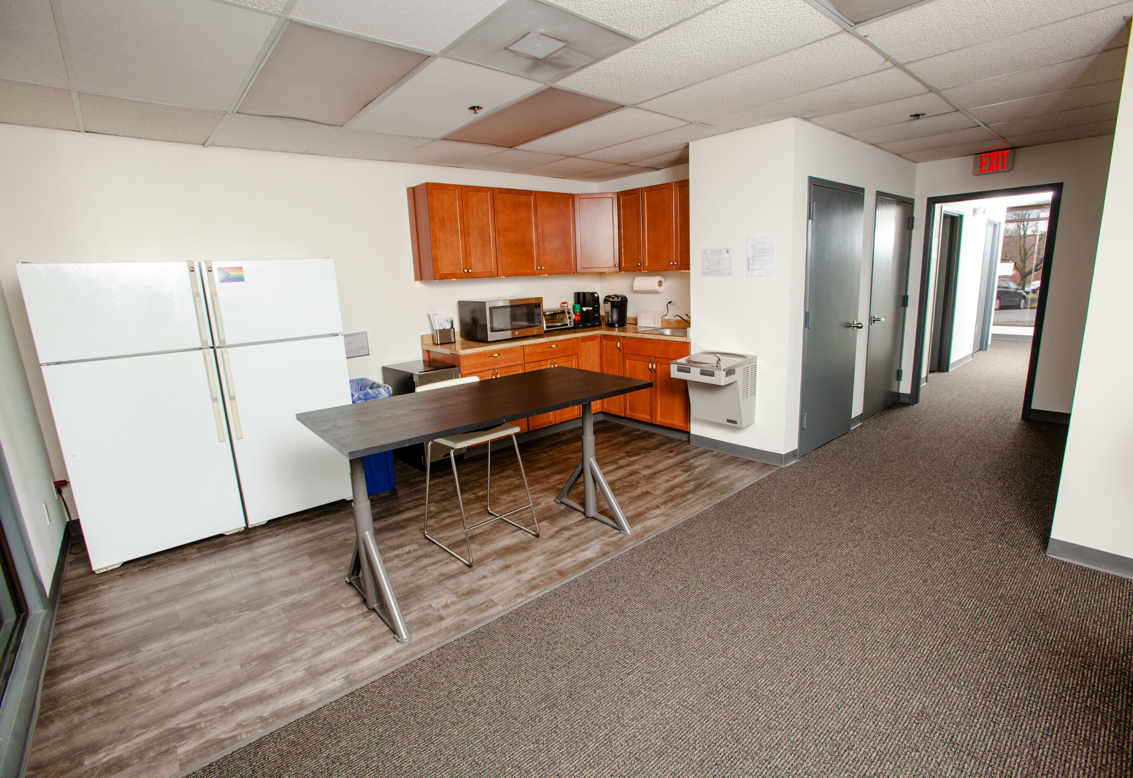 A kitchen area with wooden cabinets, a large refrigerator, a microwave, and a counter with two bar stools, adjacent to a hallway with carpeted flooring.