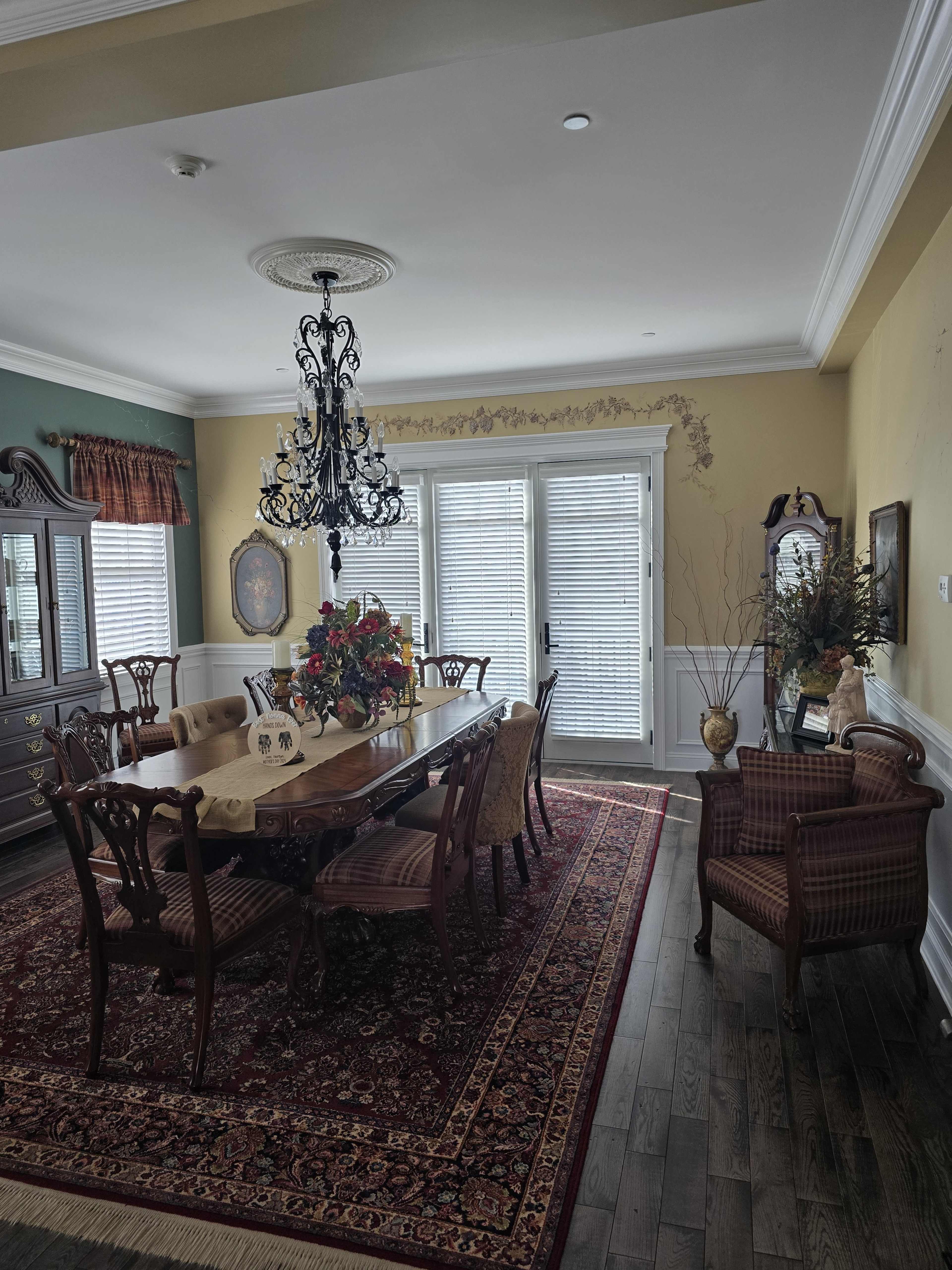 The image shows an elegant dining room featuring a large wooden table surrounded by upholstered chairs, a chandelier above, and a floral centerpiece, with decorative elements against a yellow and white wall.