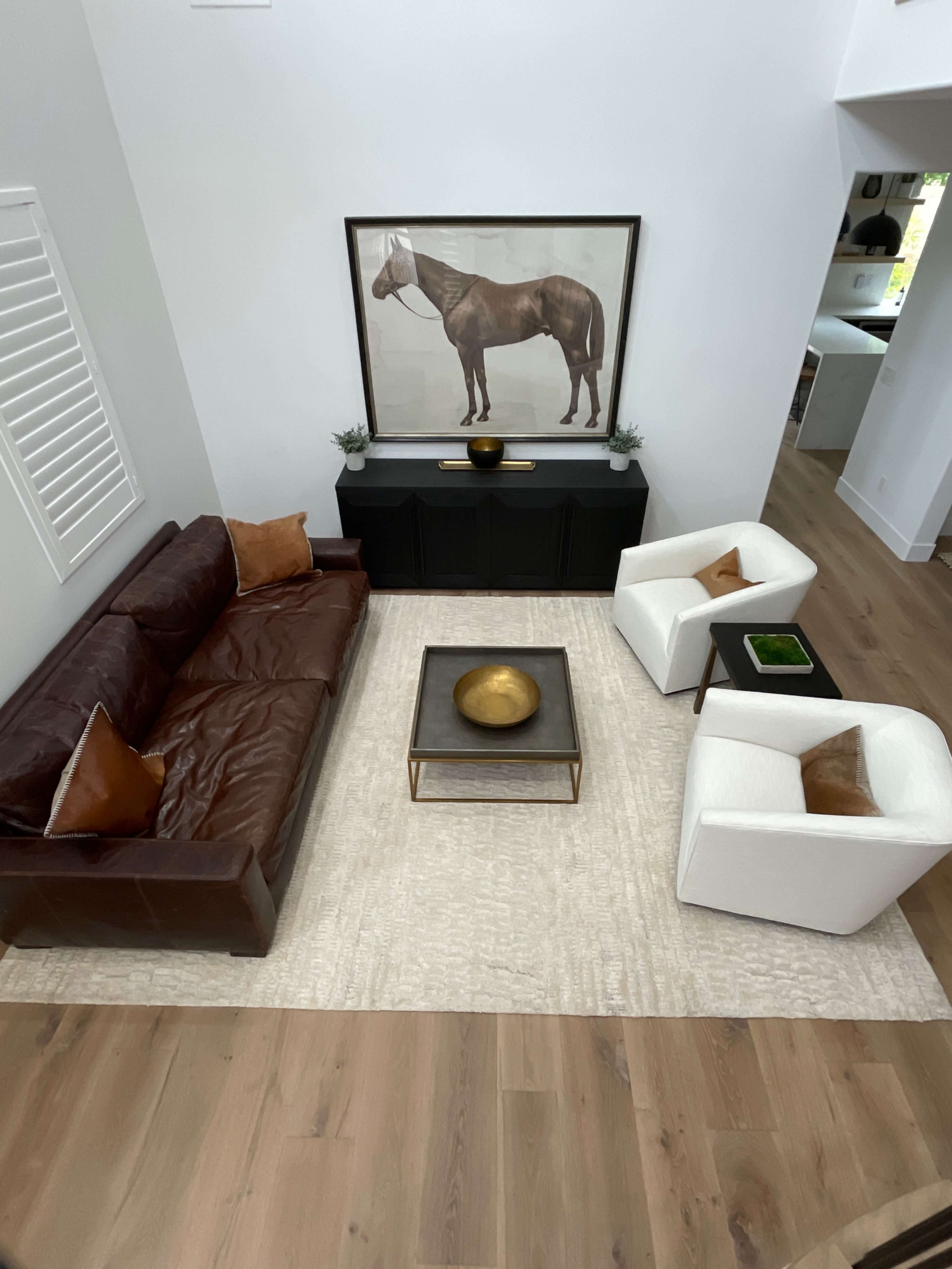 A modern living room with a brown leather sofa, two white armchairs, a coffee table with a gold bowl, and a large horse painting hanging above a dark cabinet.