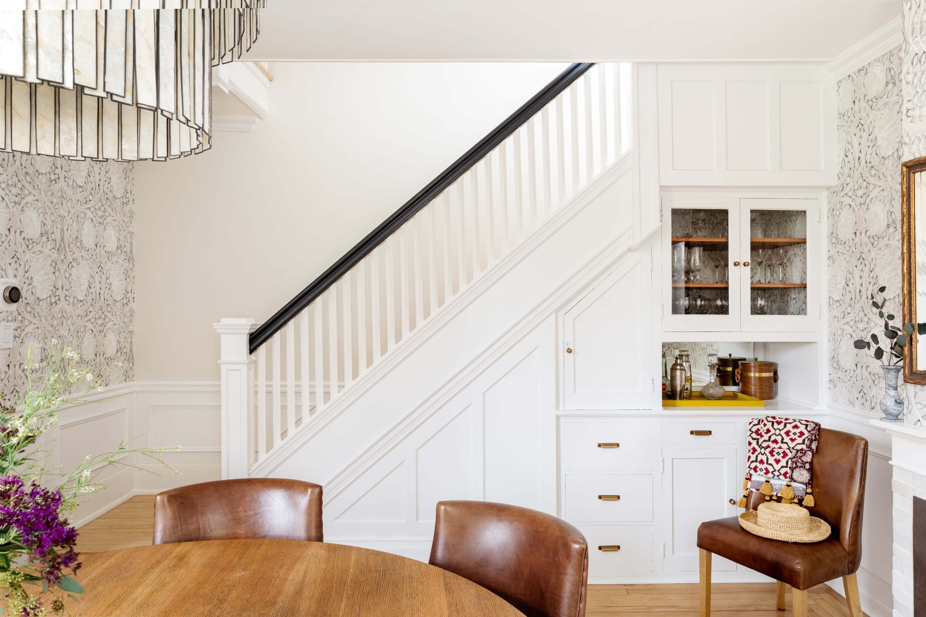 A dining area features a round wooden table with leather chairs, adjacent to a staircase leading to an upper level, with a wall decorated in patterned wallpaper and a built-in cabinet displaying glassware.