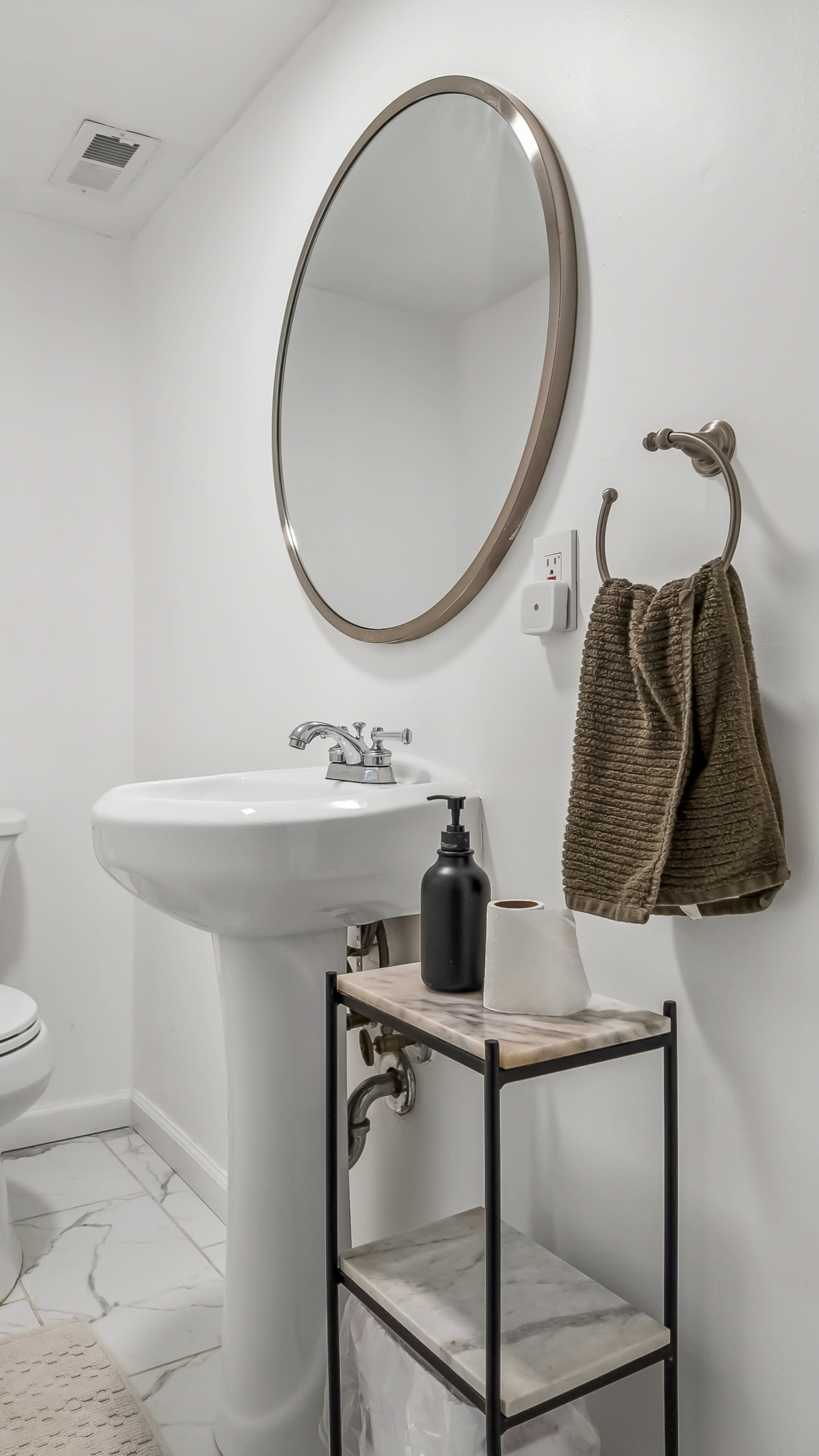 The scene features a minimalist bathroom with a round mirror above a white sink, a towel hanging on a wall-mounted holder, and a small black shelf displaying a soap dispenser and tissue box.