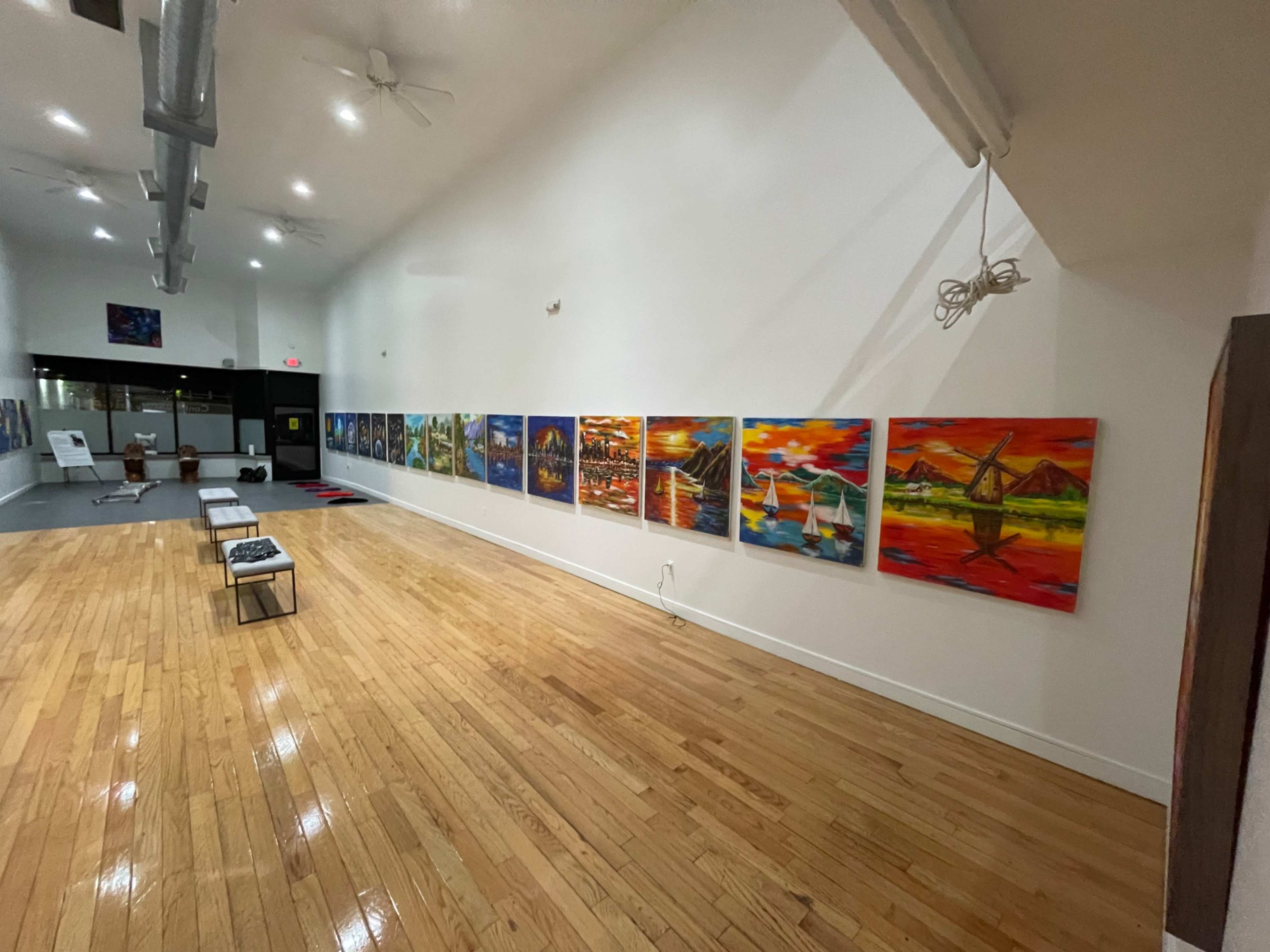 The image shows a spacious indoor gallery with polished wooden floors and a collection of colorful paintings displayed along one wall.