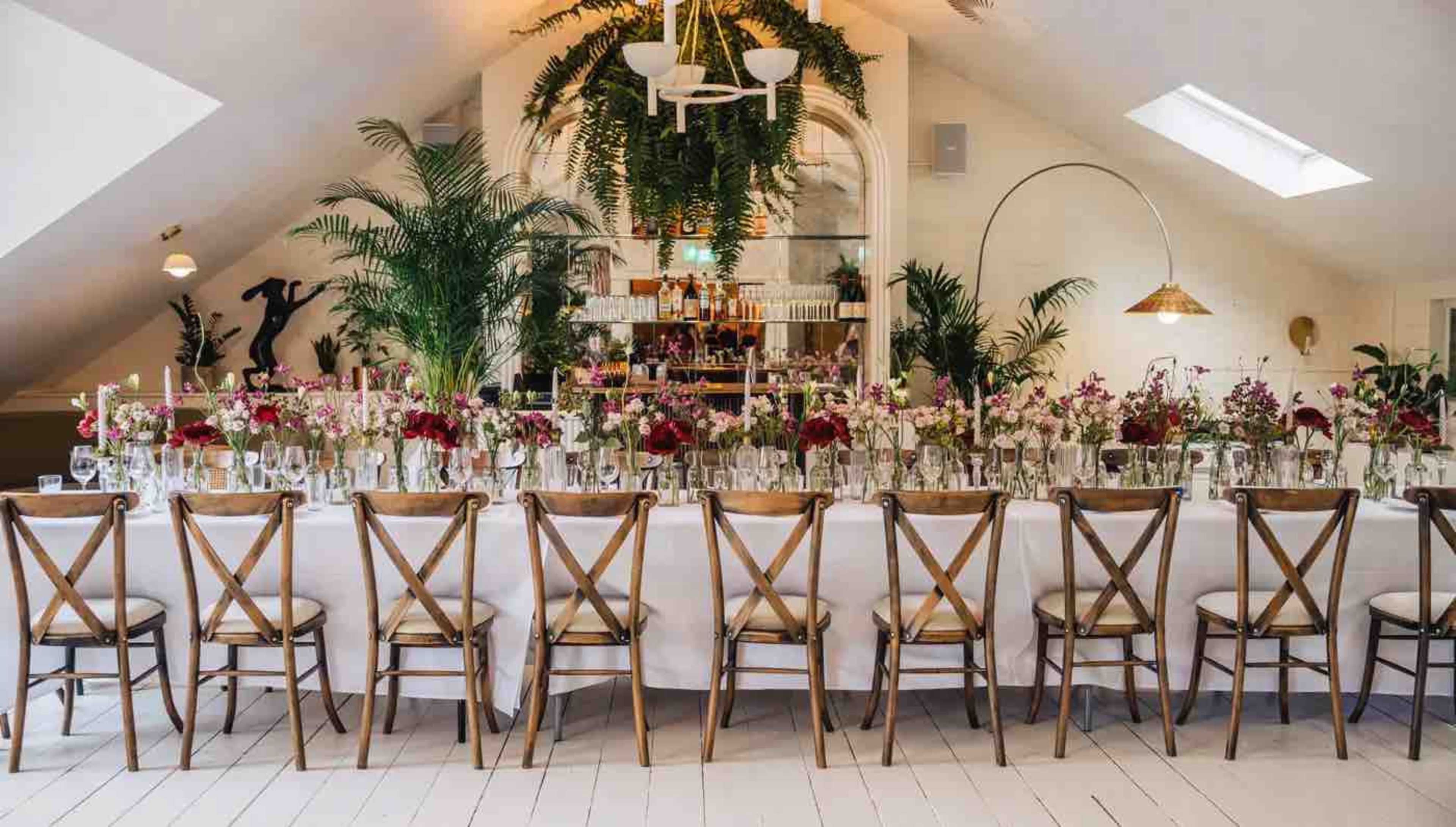 A long, elegantly arranged dining table is set with flowers and candles, surrounded by wooden chairs in a bright, modern space featuring greenery and a bar in the background.