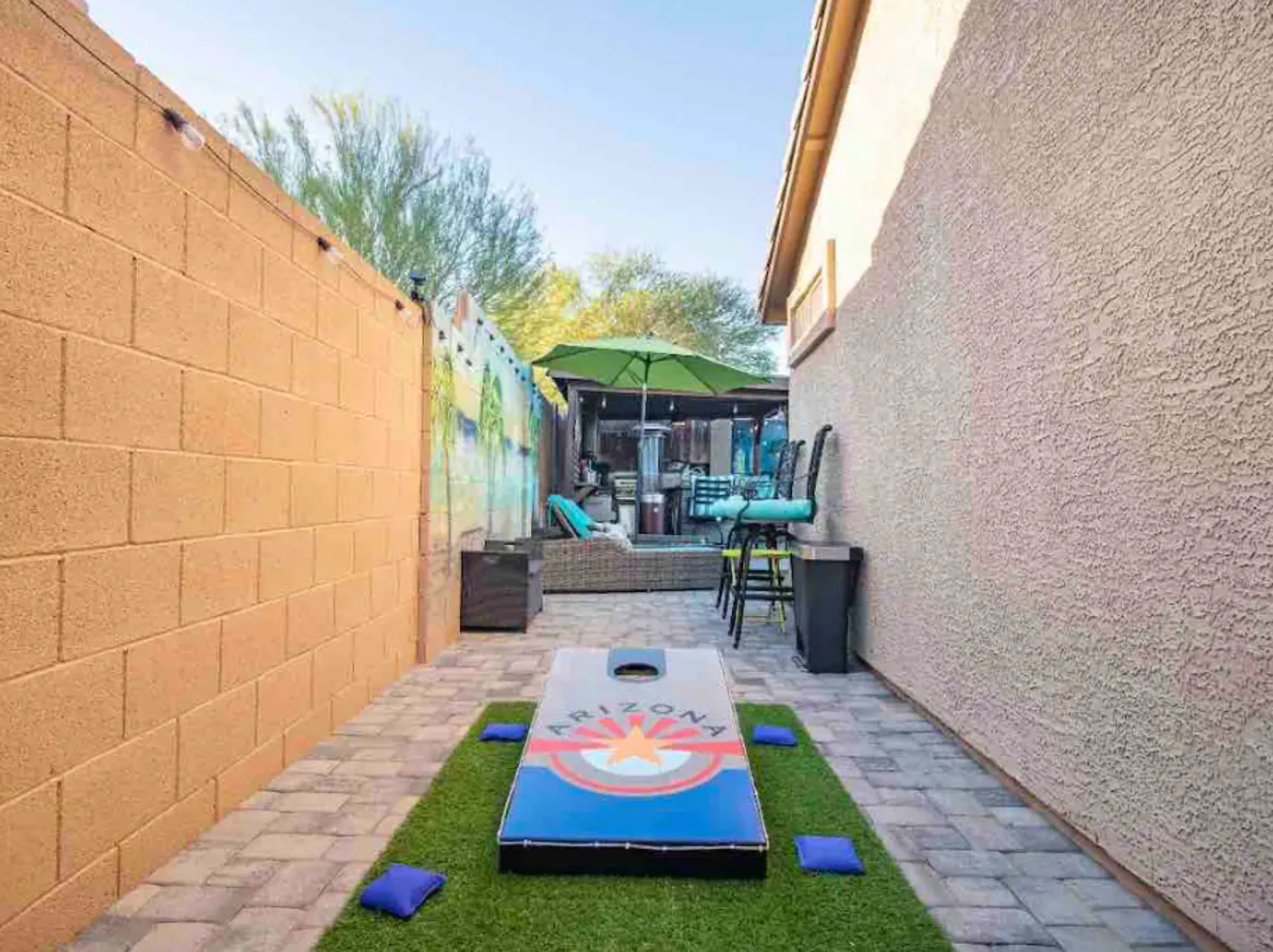 A narrow outdoor space featuring a cornhole game board on a patch of artificial grass, surrounded by walls and patio furniture under a green umbrella.