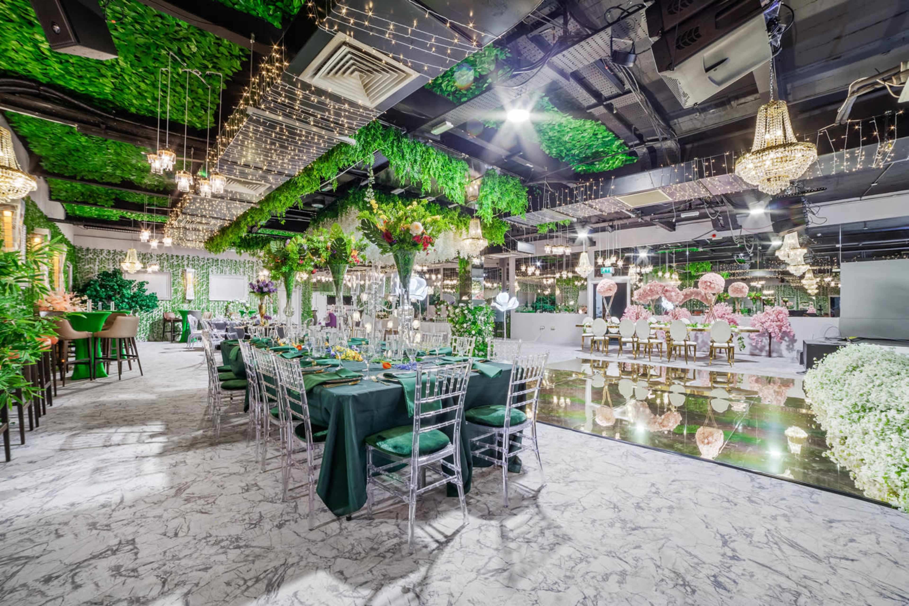 An elegantly decorated banquet hall with a long green-themed dining table, surrounded by lush greenery and floral arrangements, and illuminated by sparkling chandeliers.