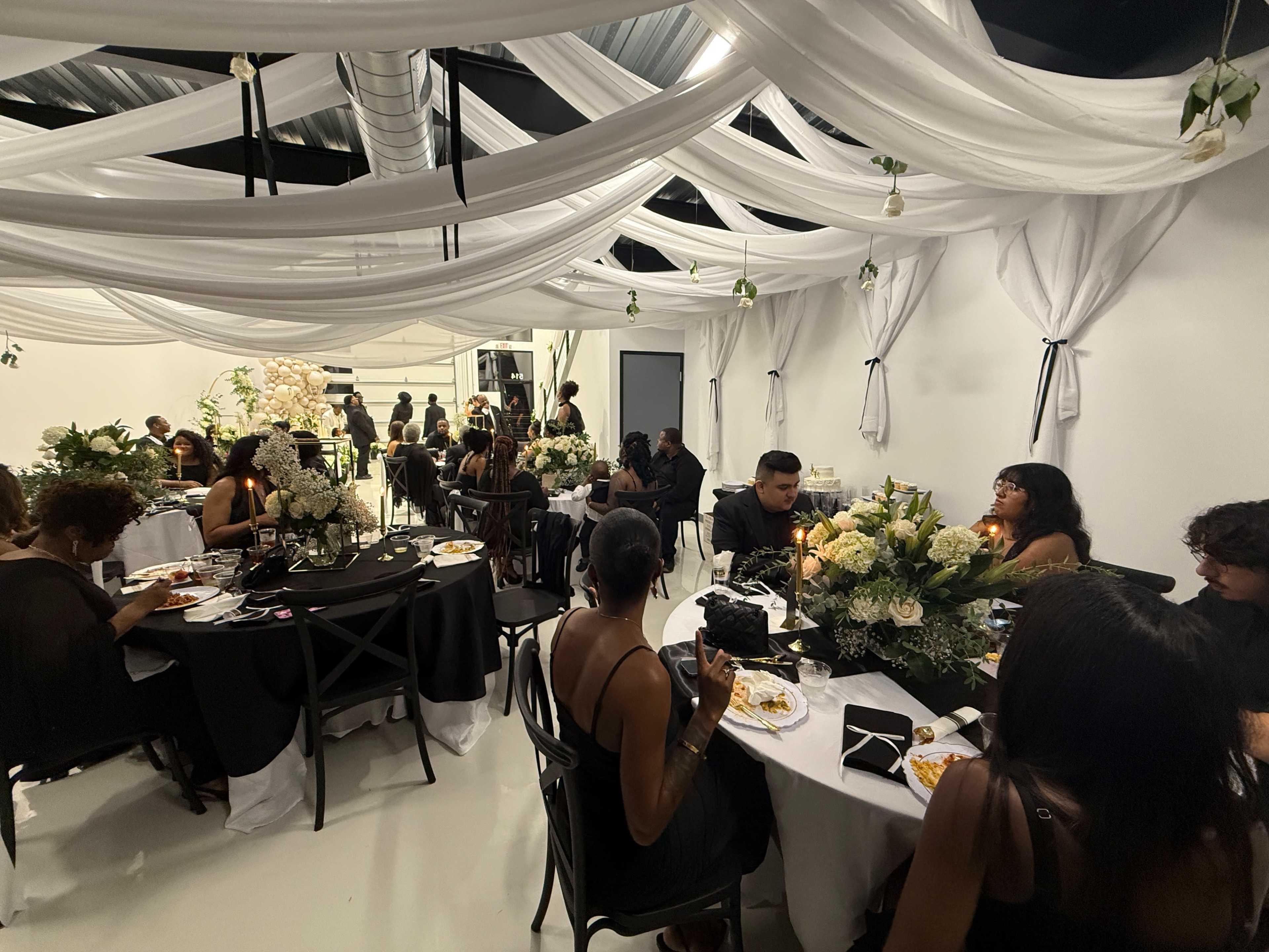 The image shows a gathering in a stylishly decorated room with guests seated at tables, dining amidst draped fabric and floral arrangements.