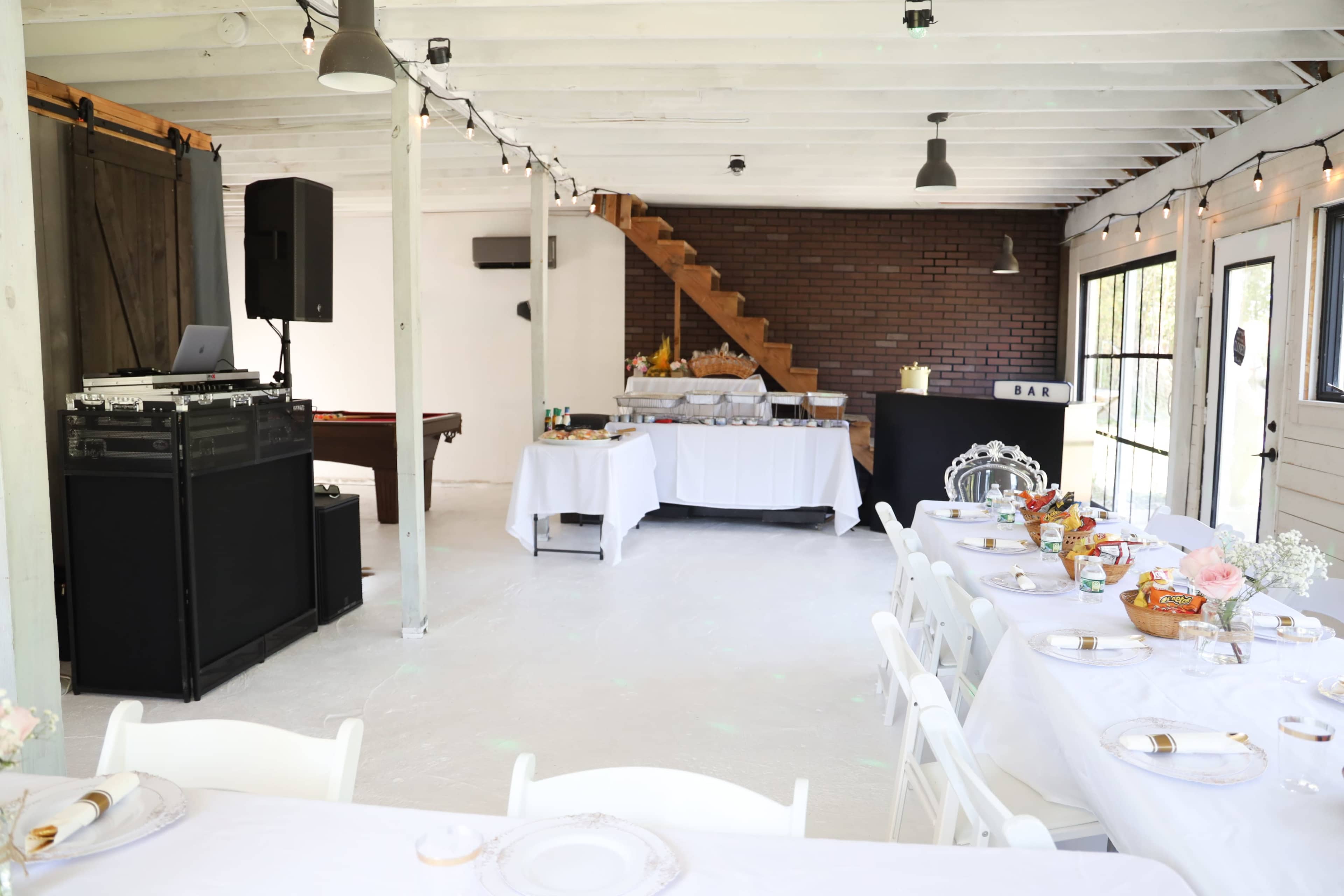 The image shows a spacious event room with a dining setup, featuring a long table covered in white tablecloths and a catering area with food on display.