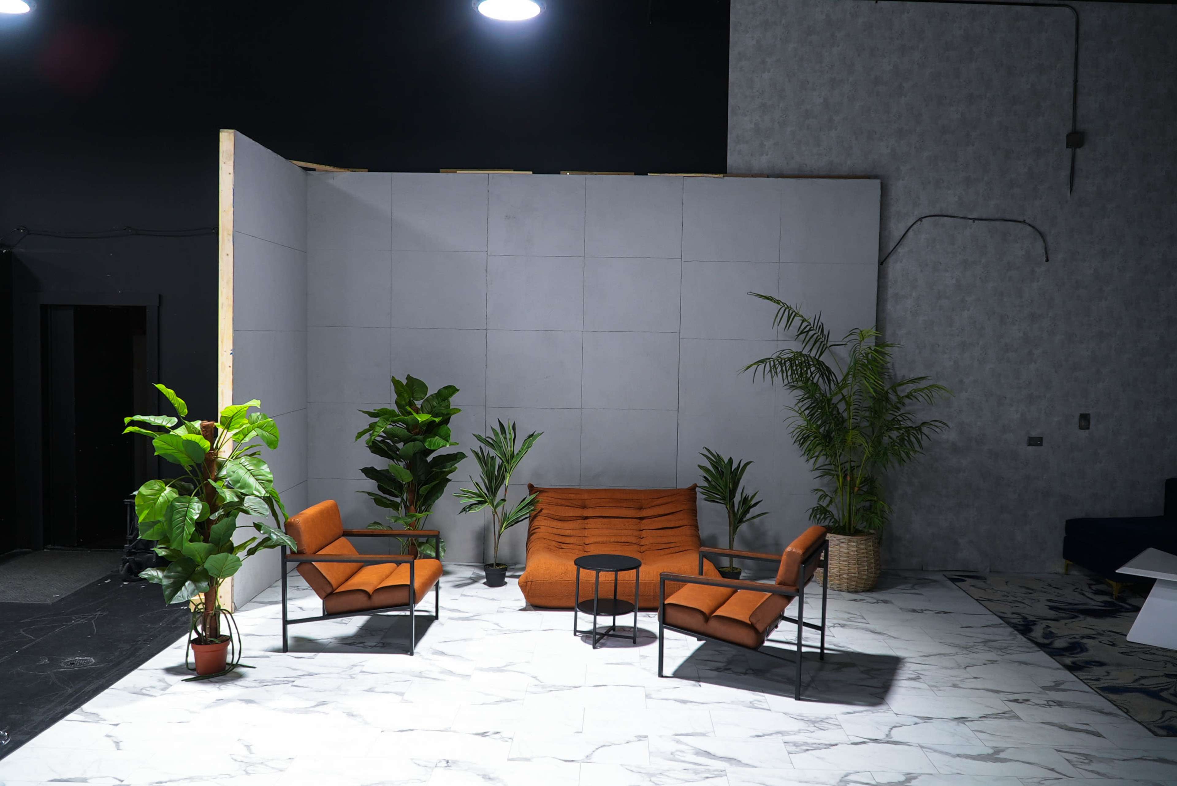 The image shows a minimalist interior setup featuring two orange chairs, a small black table, and plants arranged on a marble floor against a gray backdrop.