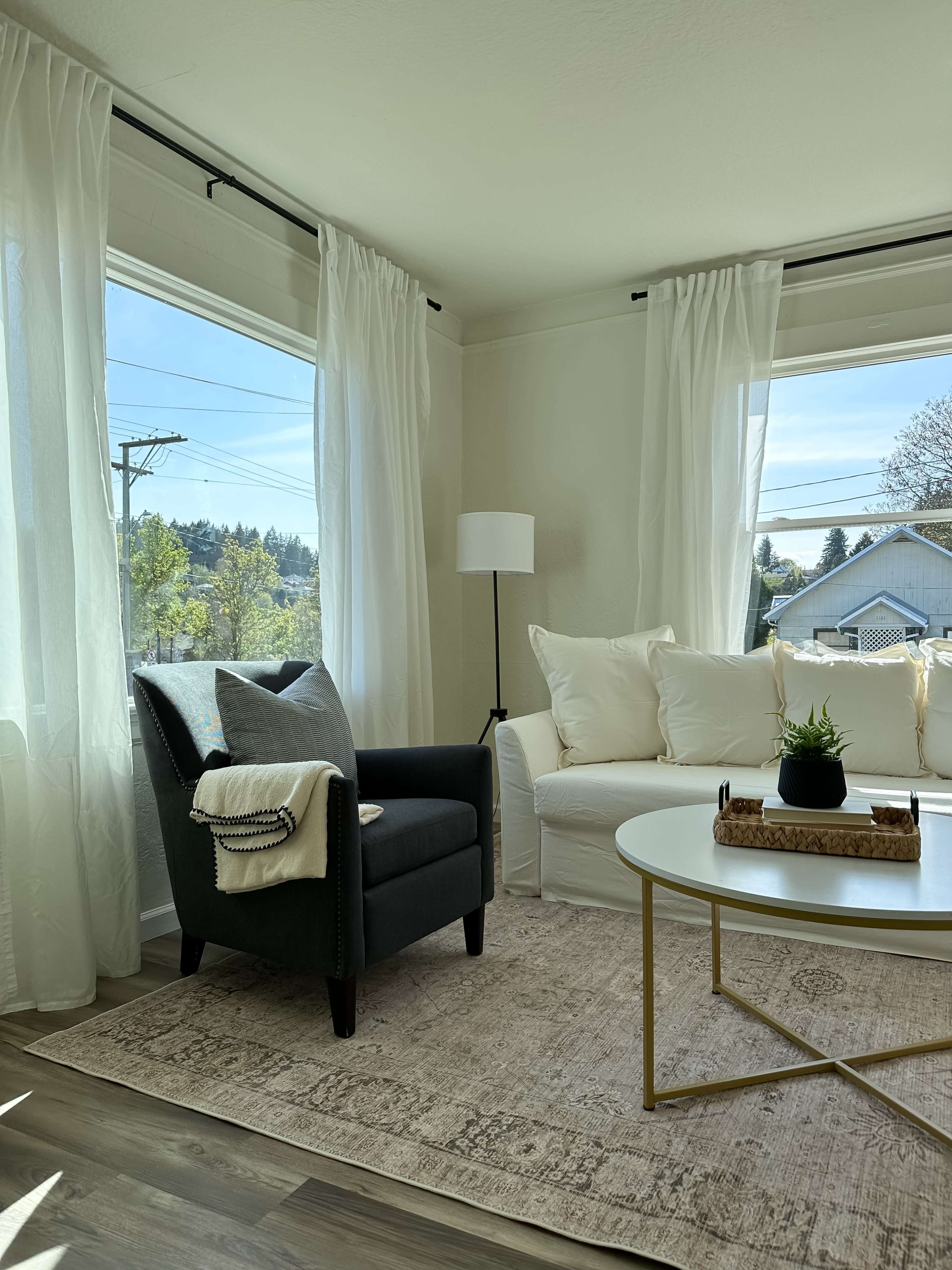 A modern living room features a gray armchair beside a white sofa, with large windows dressed in sheer curtains and a round coffee table centered on a light area rug.