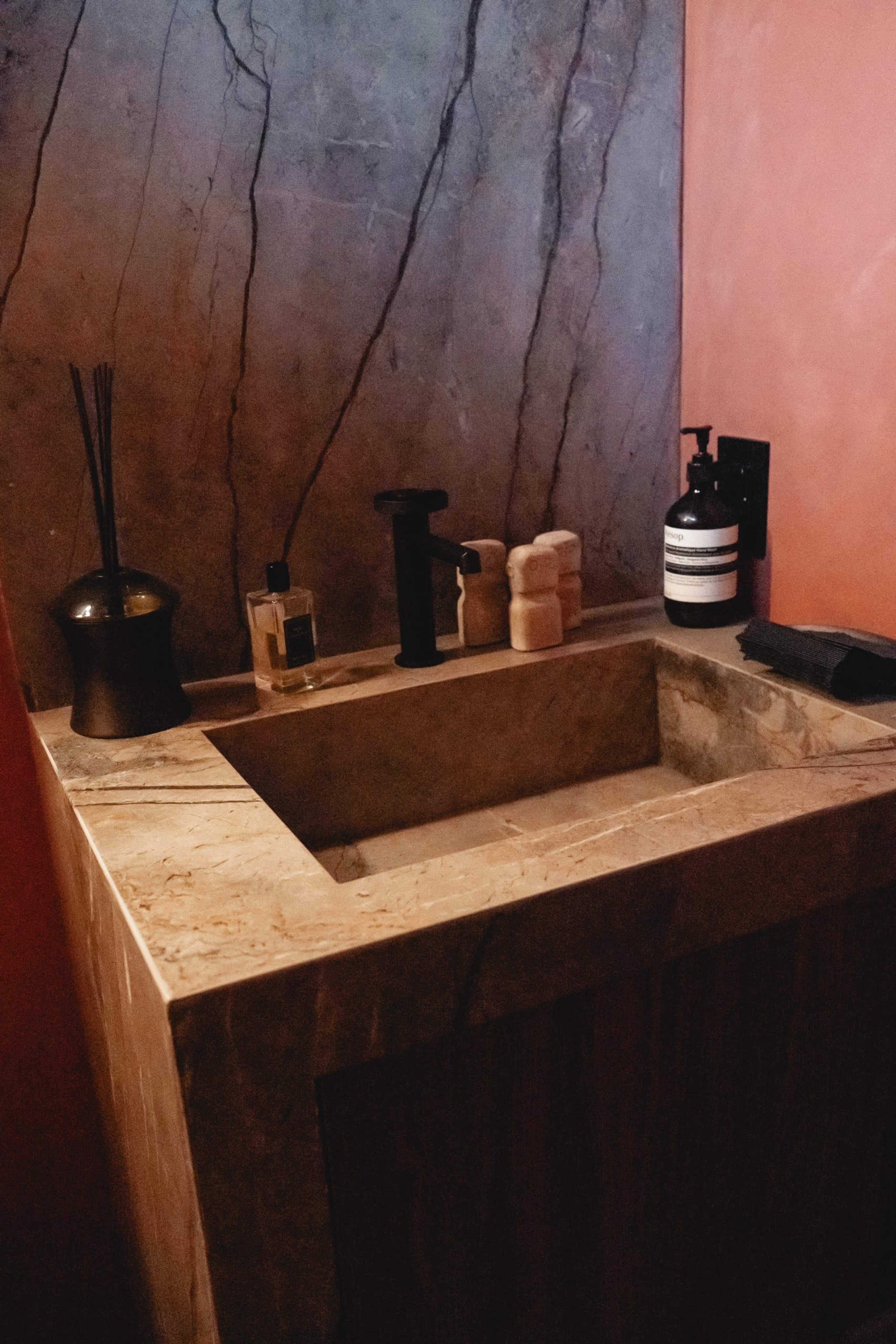 The image shows a minimalist bathroom sink area featuring a stone countertop and wall with a subtle dark pattern, accompanied by various toiletries.
