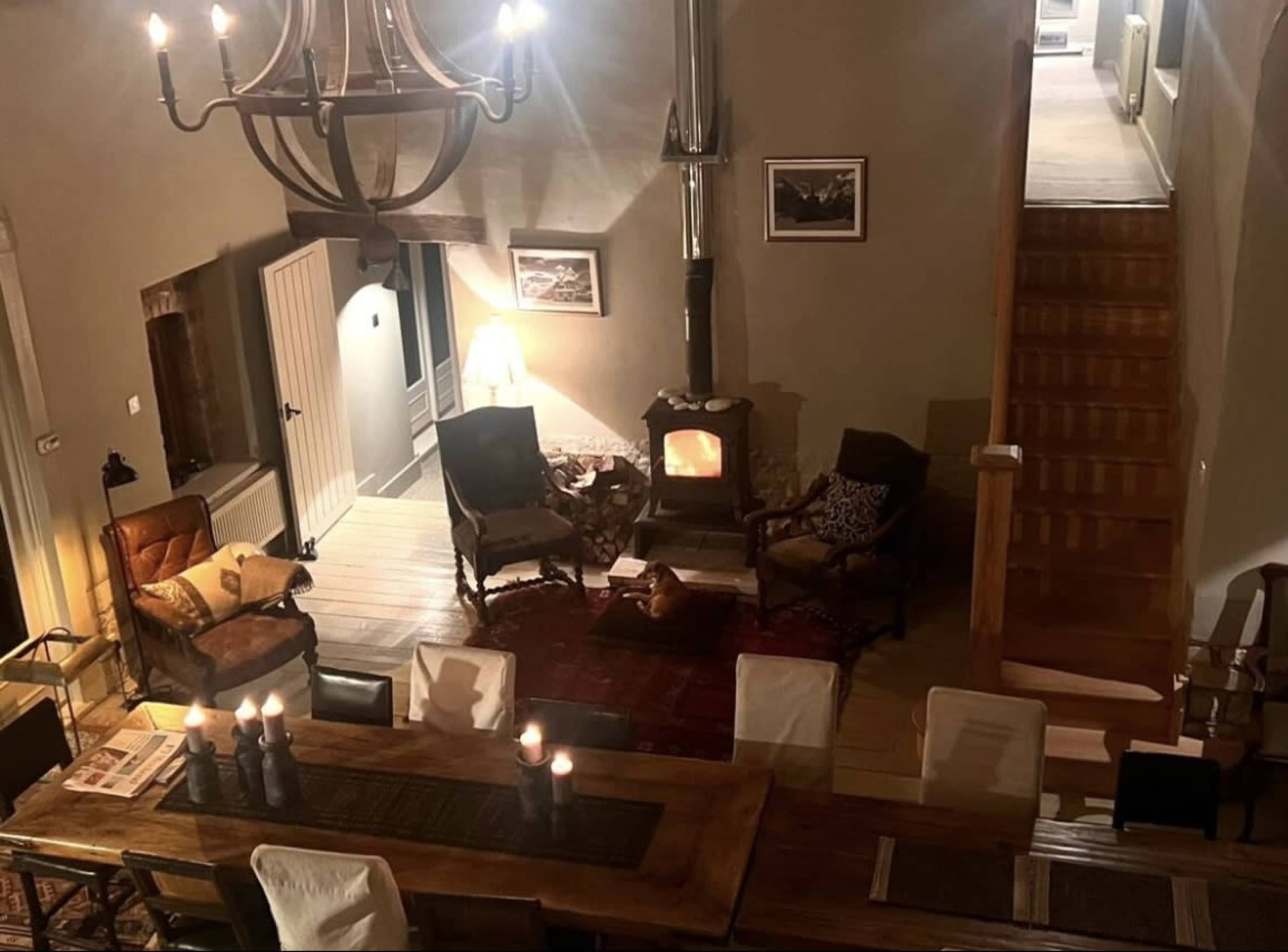 A cozy living room features a wood stove, seating areas with armchairs, a dining table, and stairs leading to another level.