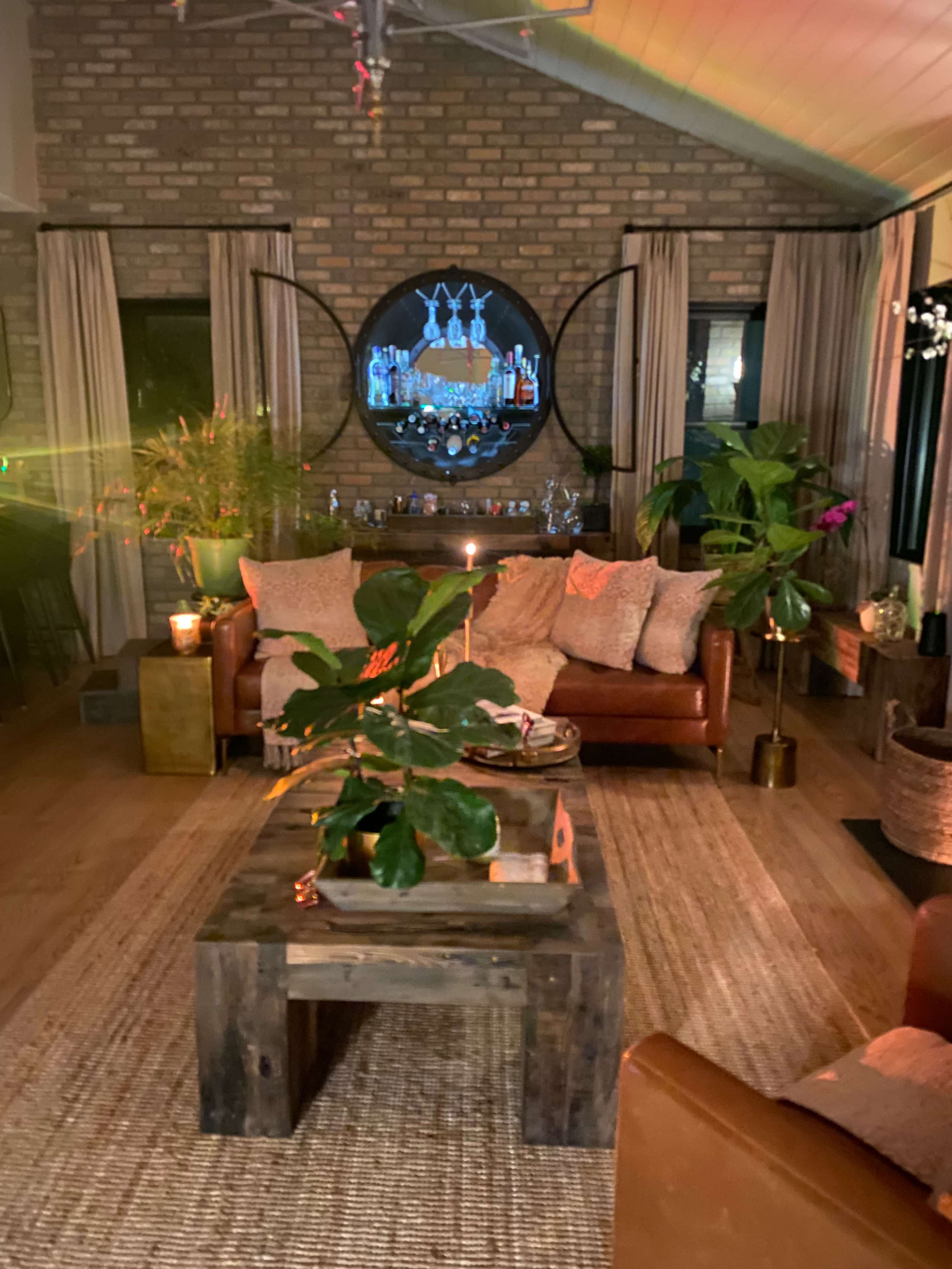 The living room features two brown leather sofas facing a large round mirror, with a wooden coffee table surrounded by plants and soft lighting.