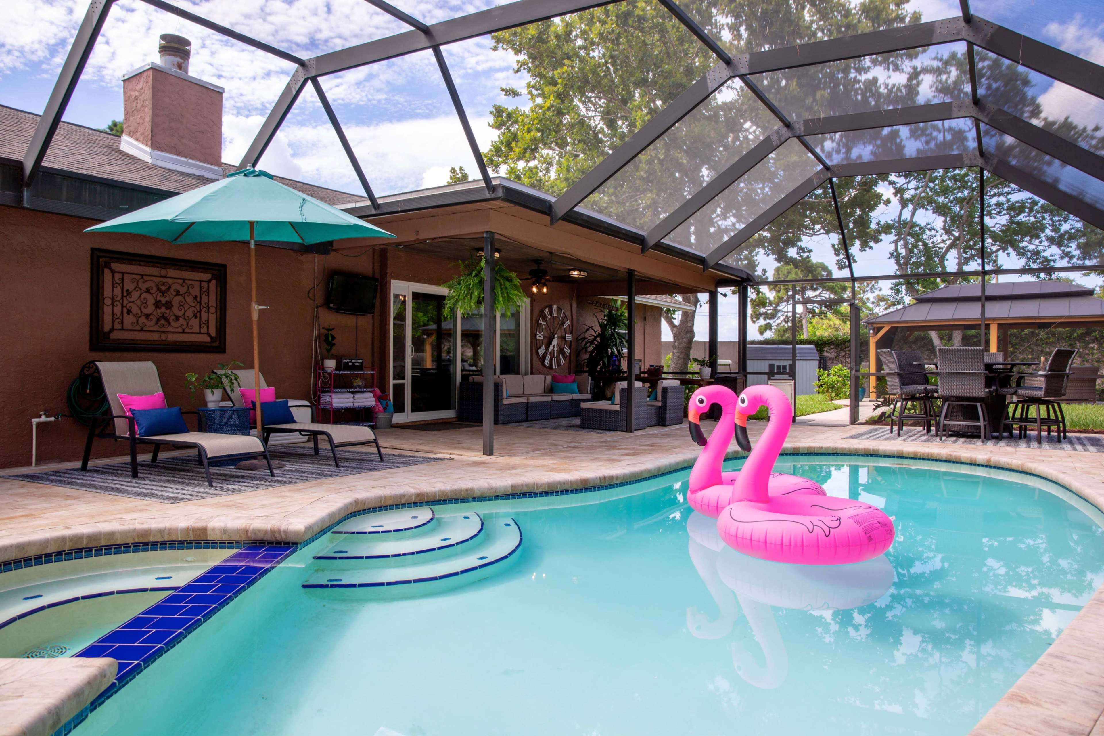 A swimming pool with a pink flamingo float is surrounded by patio furniture and a shaded area, with a house and lush greenery in the background.