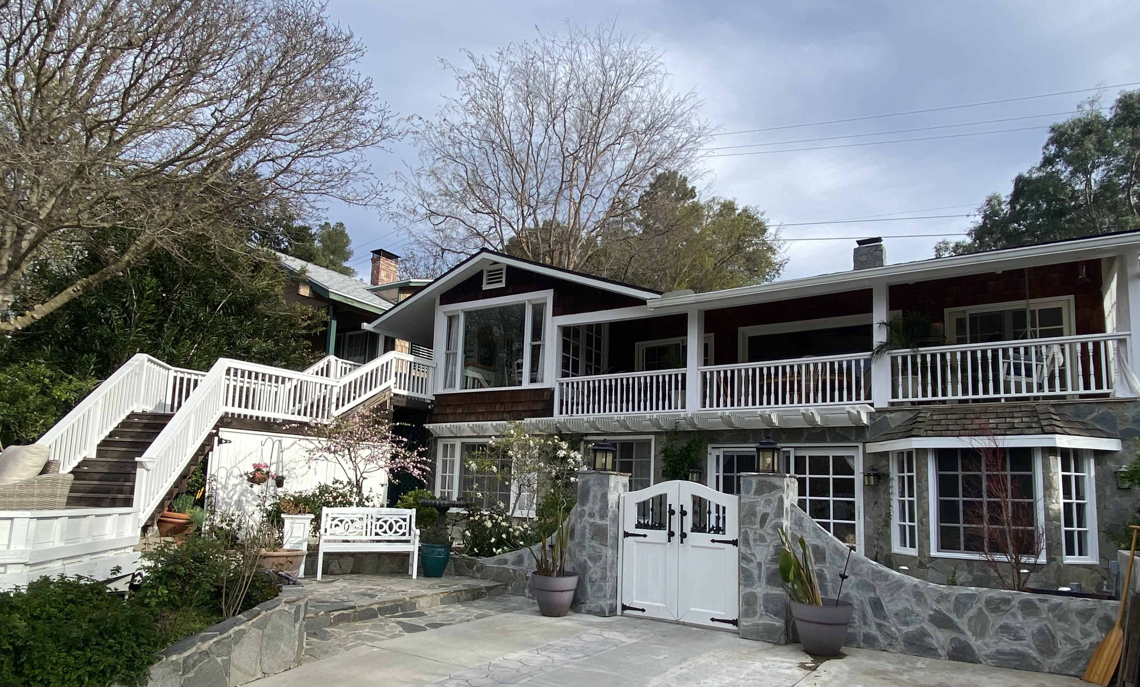 A two-story house with large balconies, stone steps, and landscaped plants, situated among trees.